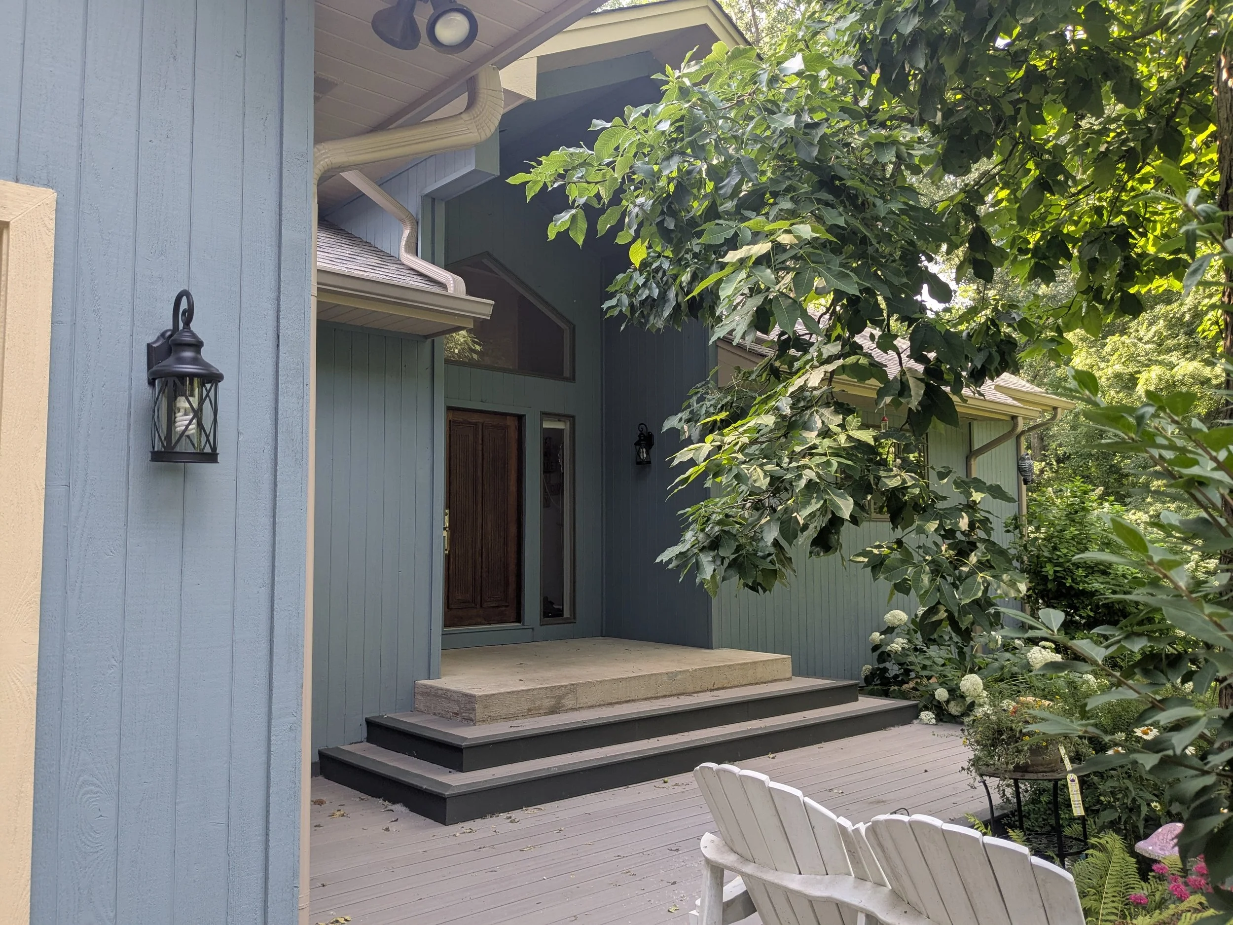 Front porch of a house with stairs, green siding, and a wooden door, surrounded by greenery including trees and flowers, with a white Adirondack chair on the porch.