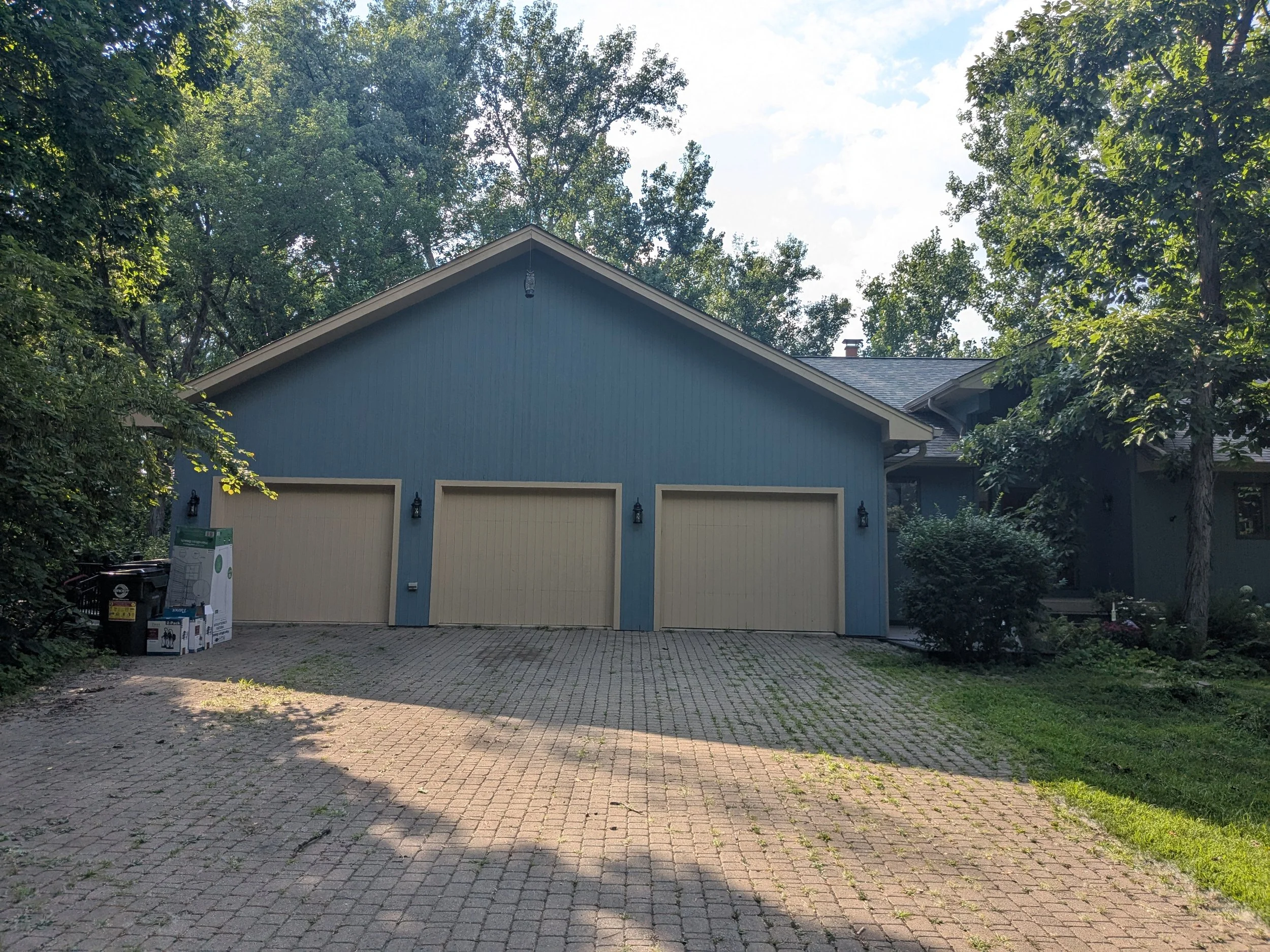 A blue house with three garage doors, surrounded by trees and bushes, in a residential area during daylight.
