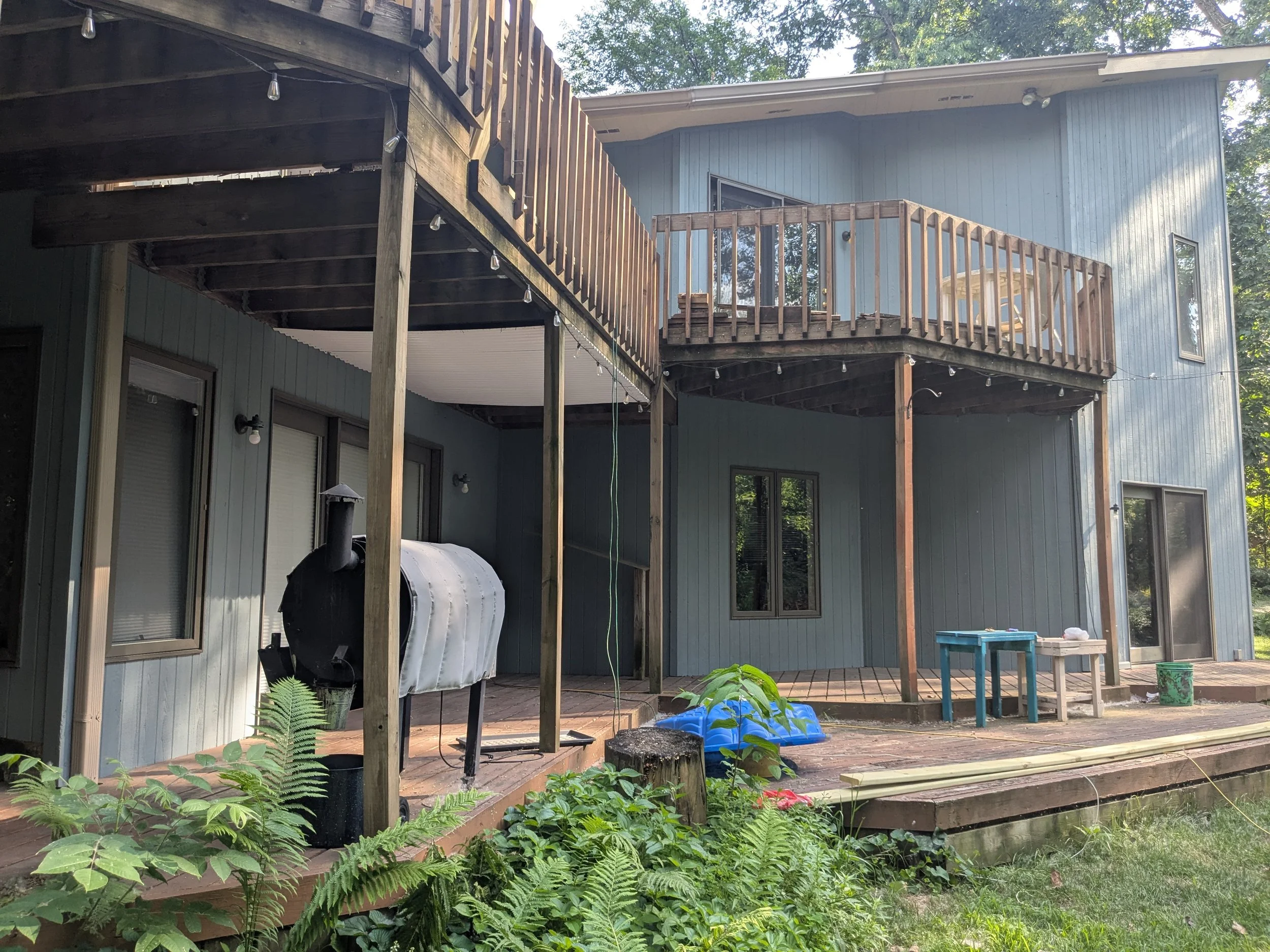 Backyard view of a two-story house with a wooden deck and railing on both levels. There is a black smoker or grill on the lower deck, with an outdoor table and some other items nearby. Green plants and ferns grow in the foreground.