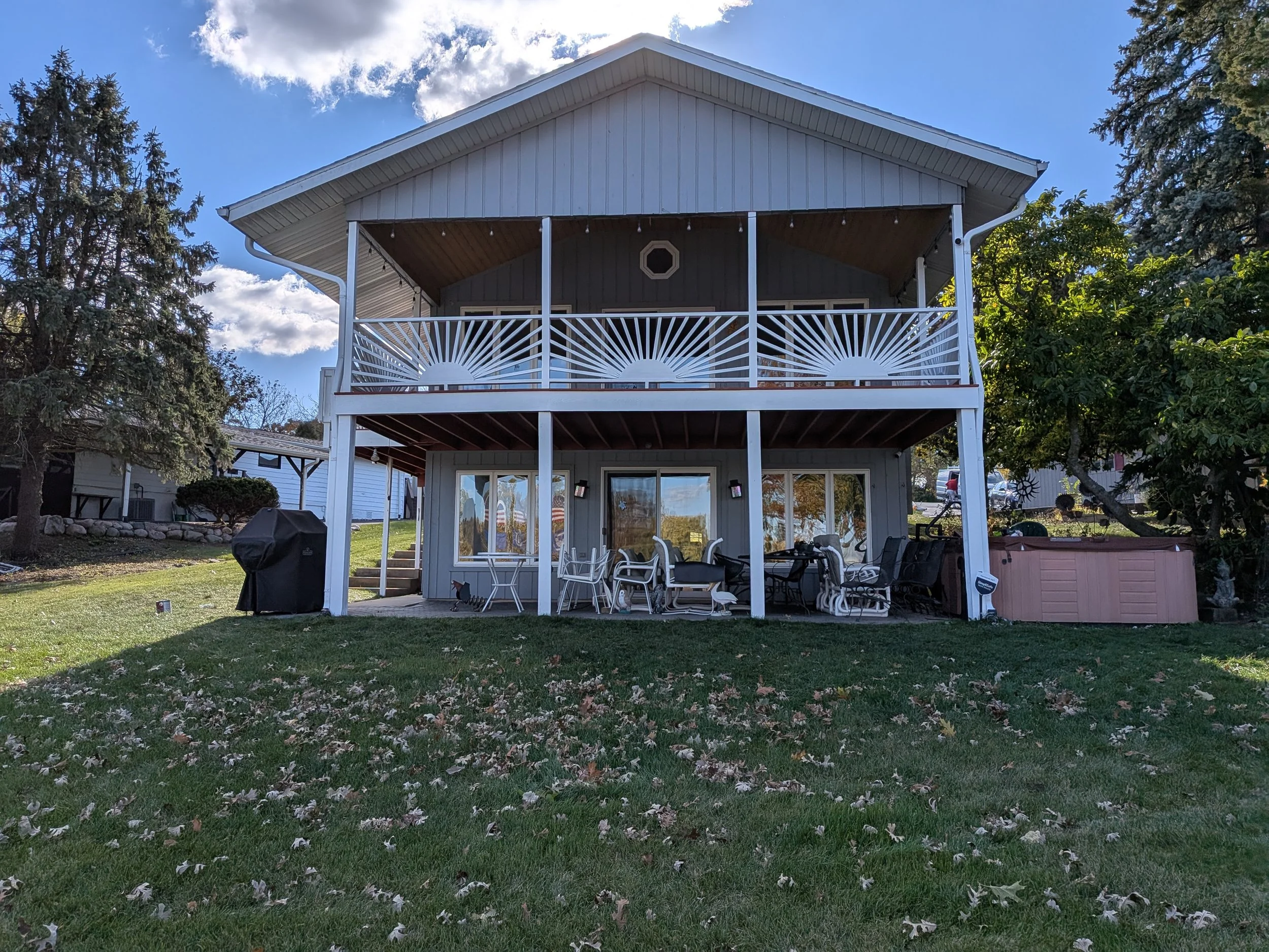 A two-story house with a white wooden exterior, a front porch on both levels, and patio furniture on the ground level. There are trees and a grassy yard with fallen leaves in the foreground. The sky is partly cloudy with sunlight.