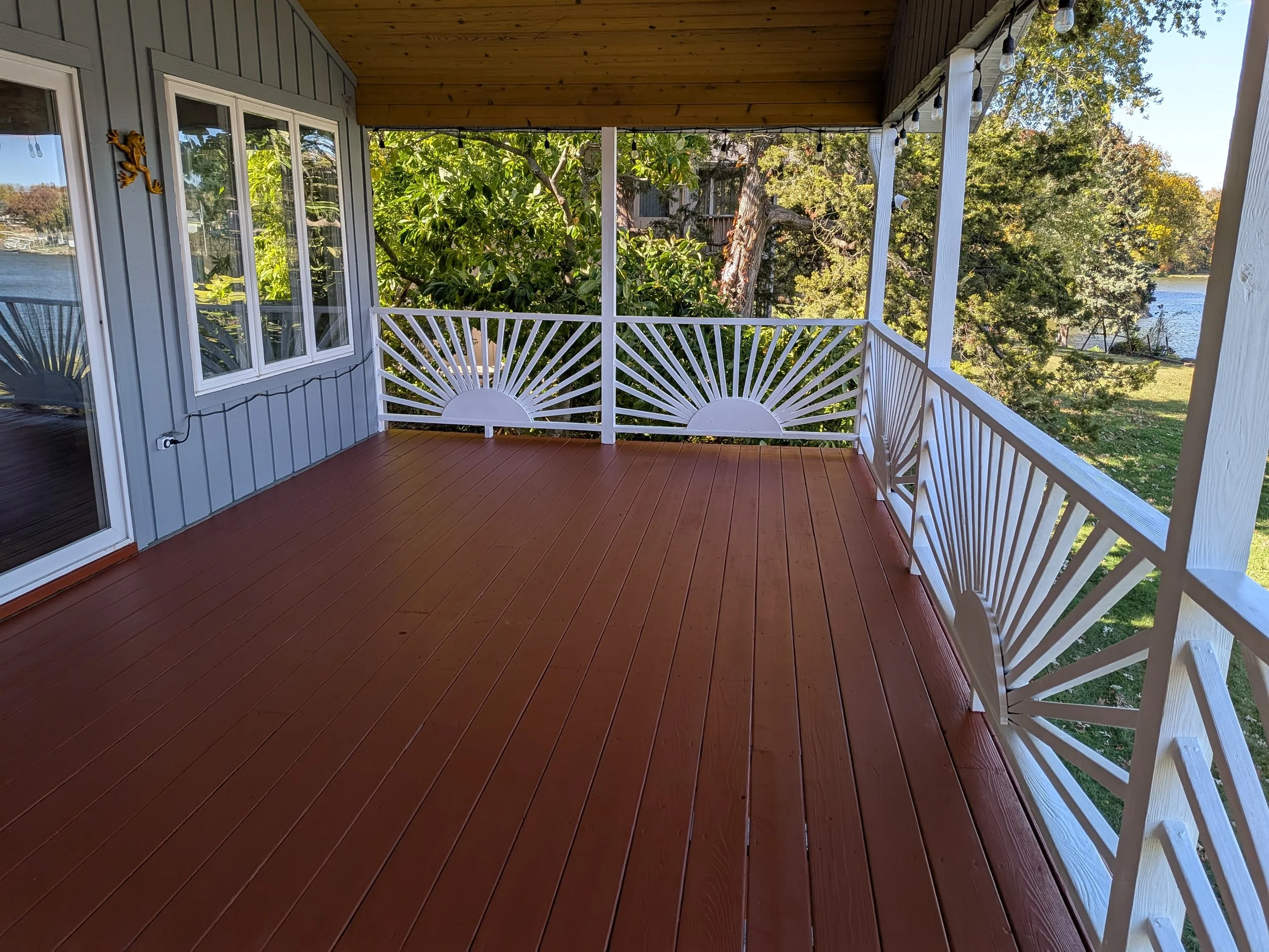 Empty wooden porch with white decorative railing overlooking a green yard and a body of water, with trees and blue sky in the background.