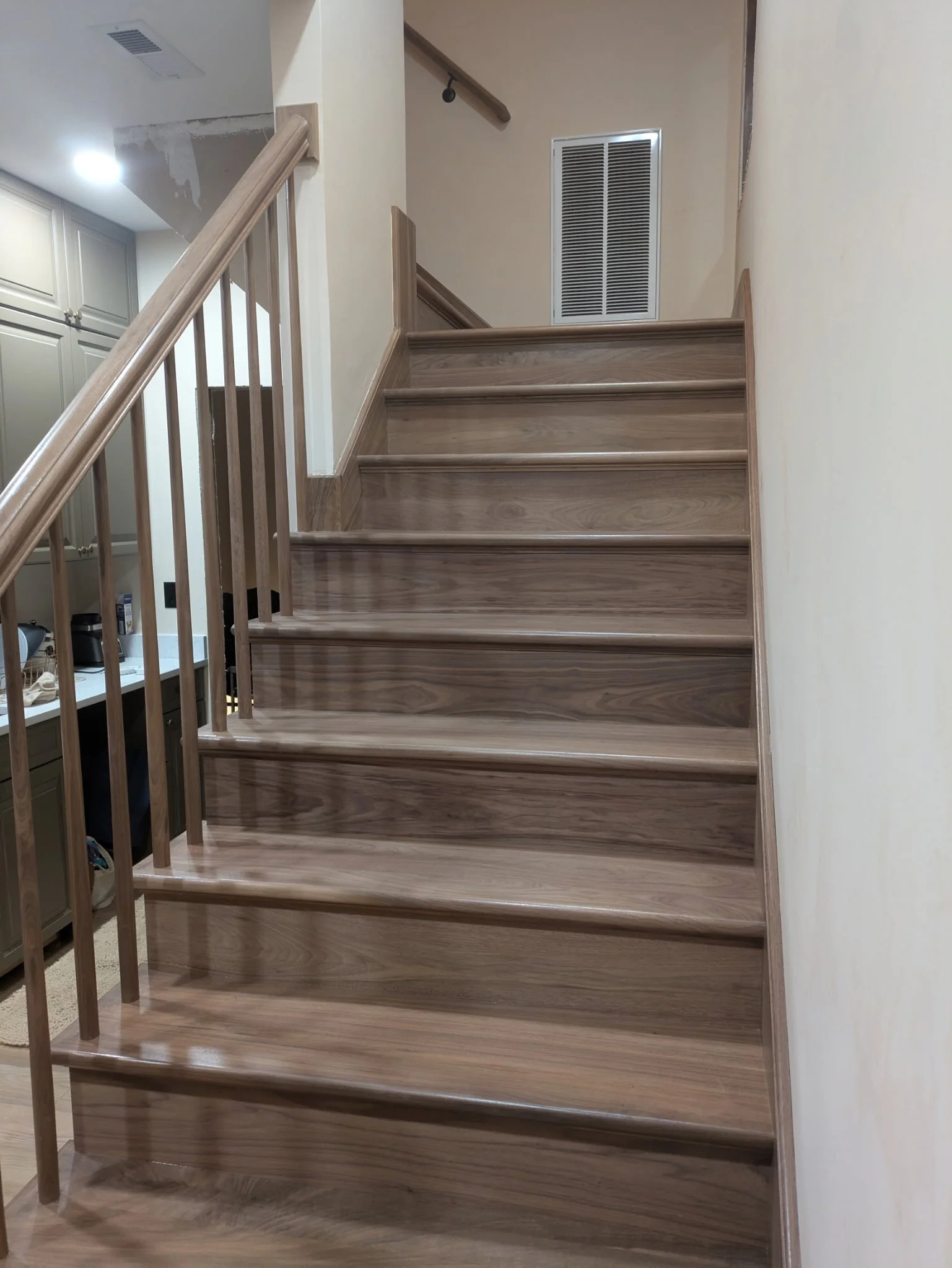 Wooden staircase with a light finish, leading up to a landing. There is a wall with an air vent at the top of the stairs and a beige wall on the right. Part of a kitchen or living area is visible on the left.