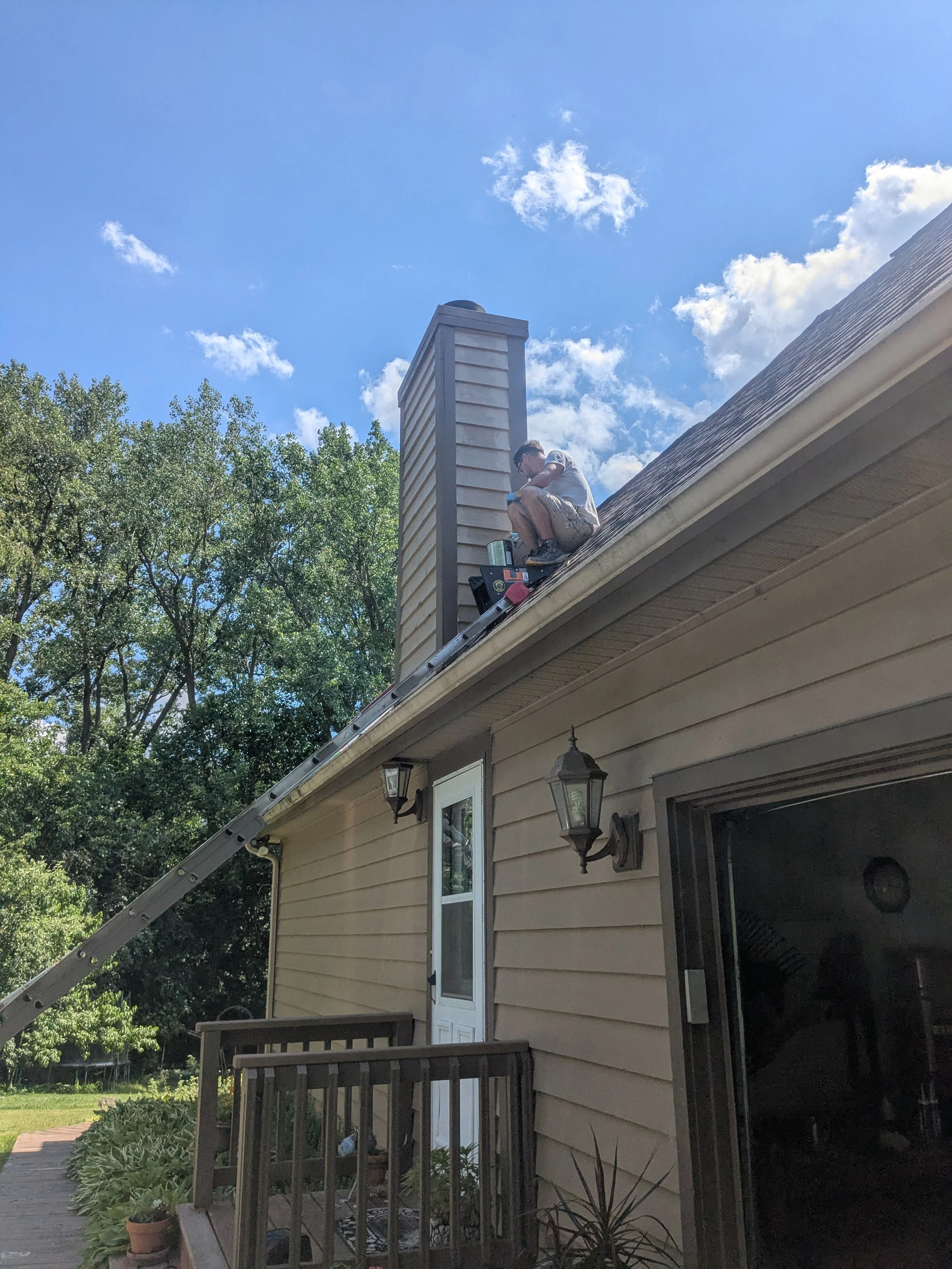A person working on a chimney on the roof of a house on a sunny day with a blue sky and some clouds, surrounded by trees.