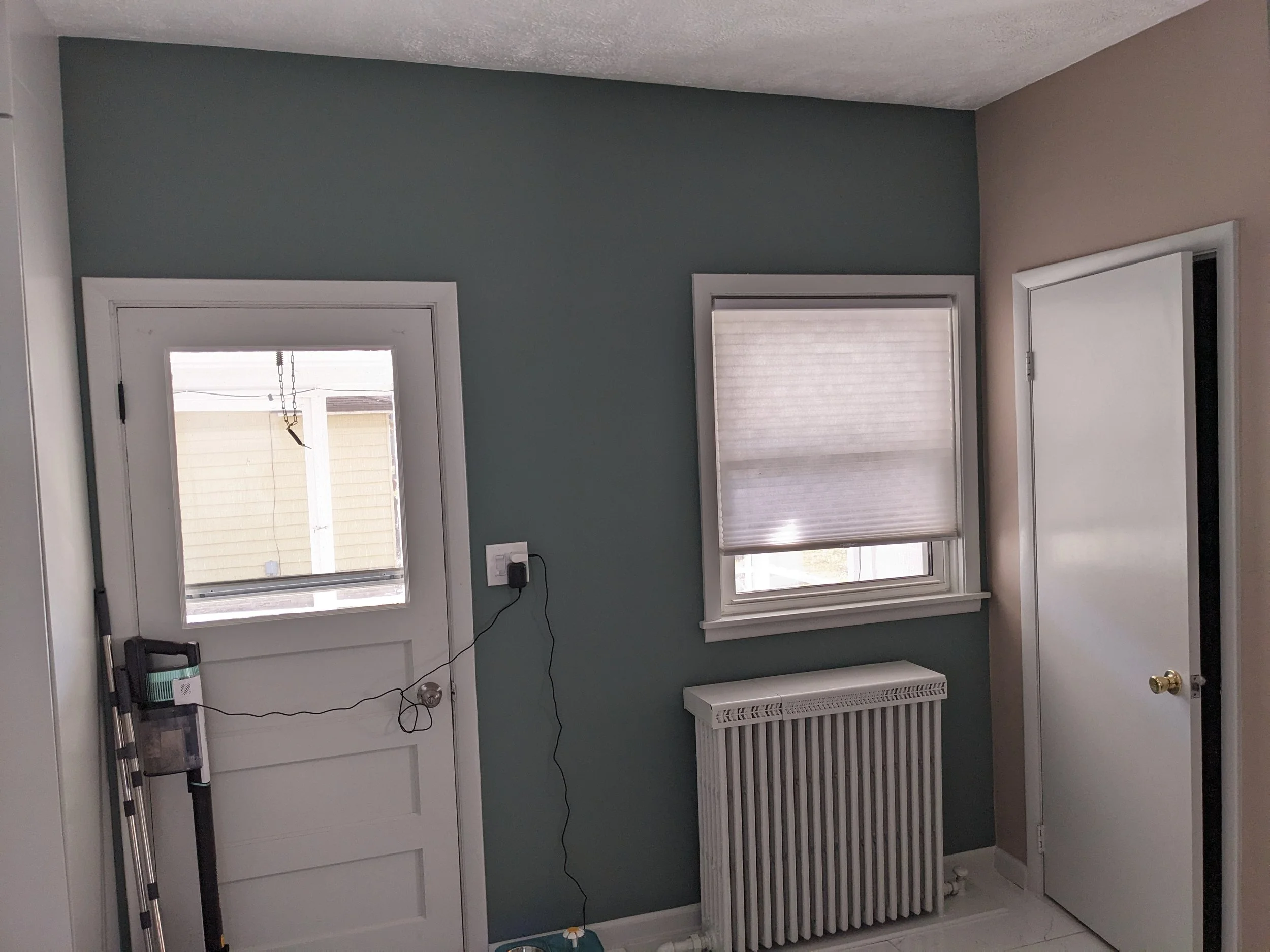 An interior room with a blue accent wall, a window with blinds, a white door with a window, a radiator, and a closed white closet door. There are also some tools leaning against the wall.
