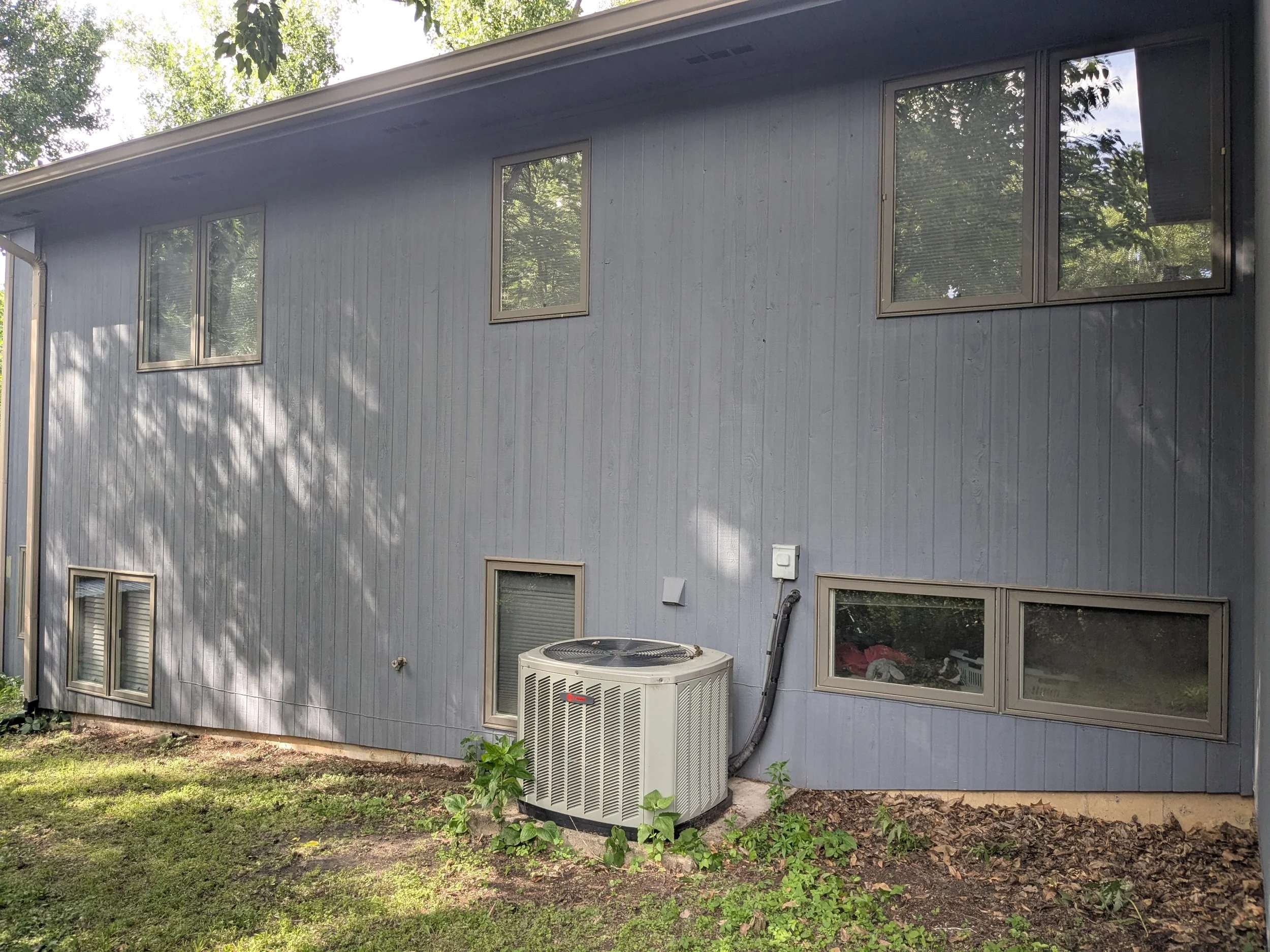 Back side of a blue house with six windows and an air conditioning unit outside, surrounded by grass and trees.