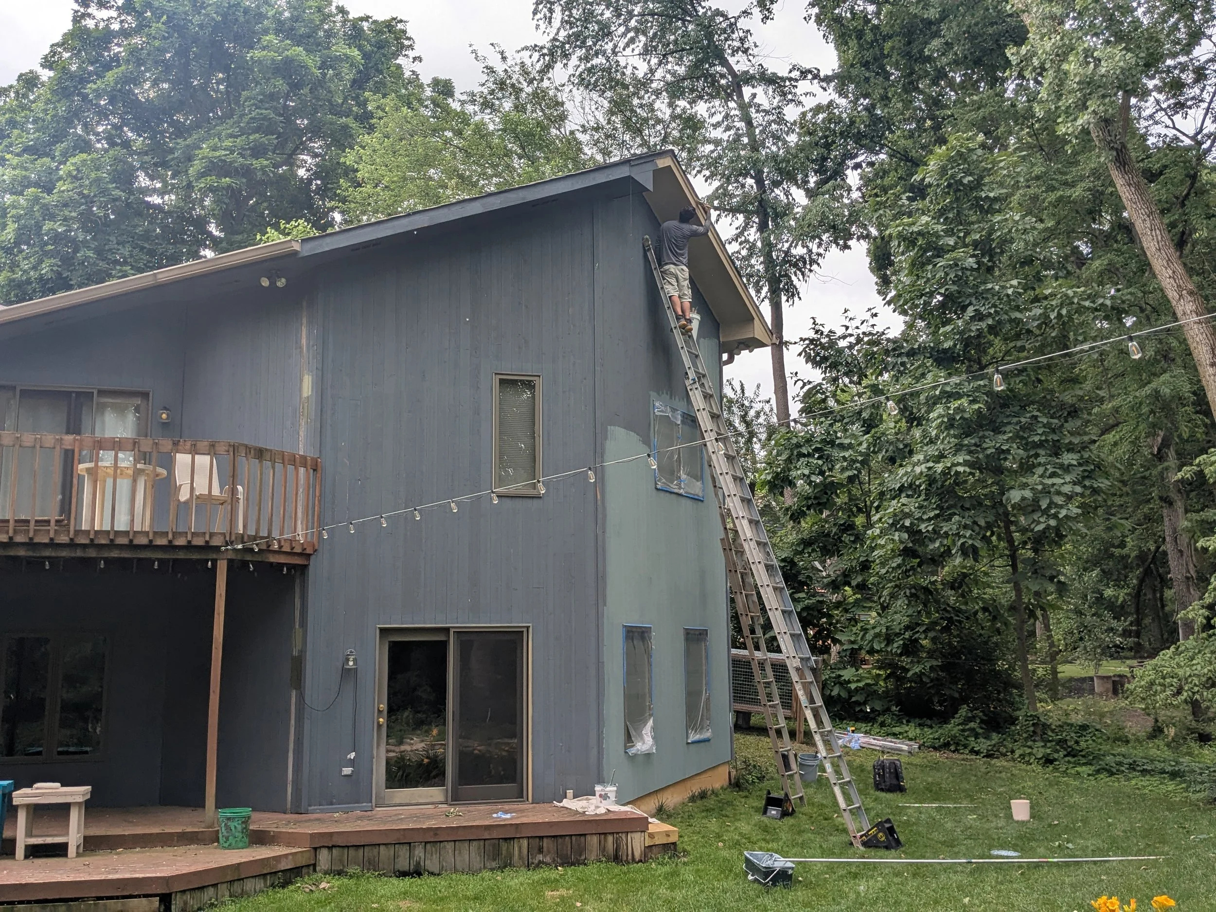 Man on a ladder working on the exterior of a two-story blue house surrounded by trees, with a deck and string lights.