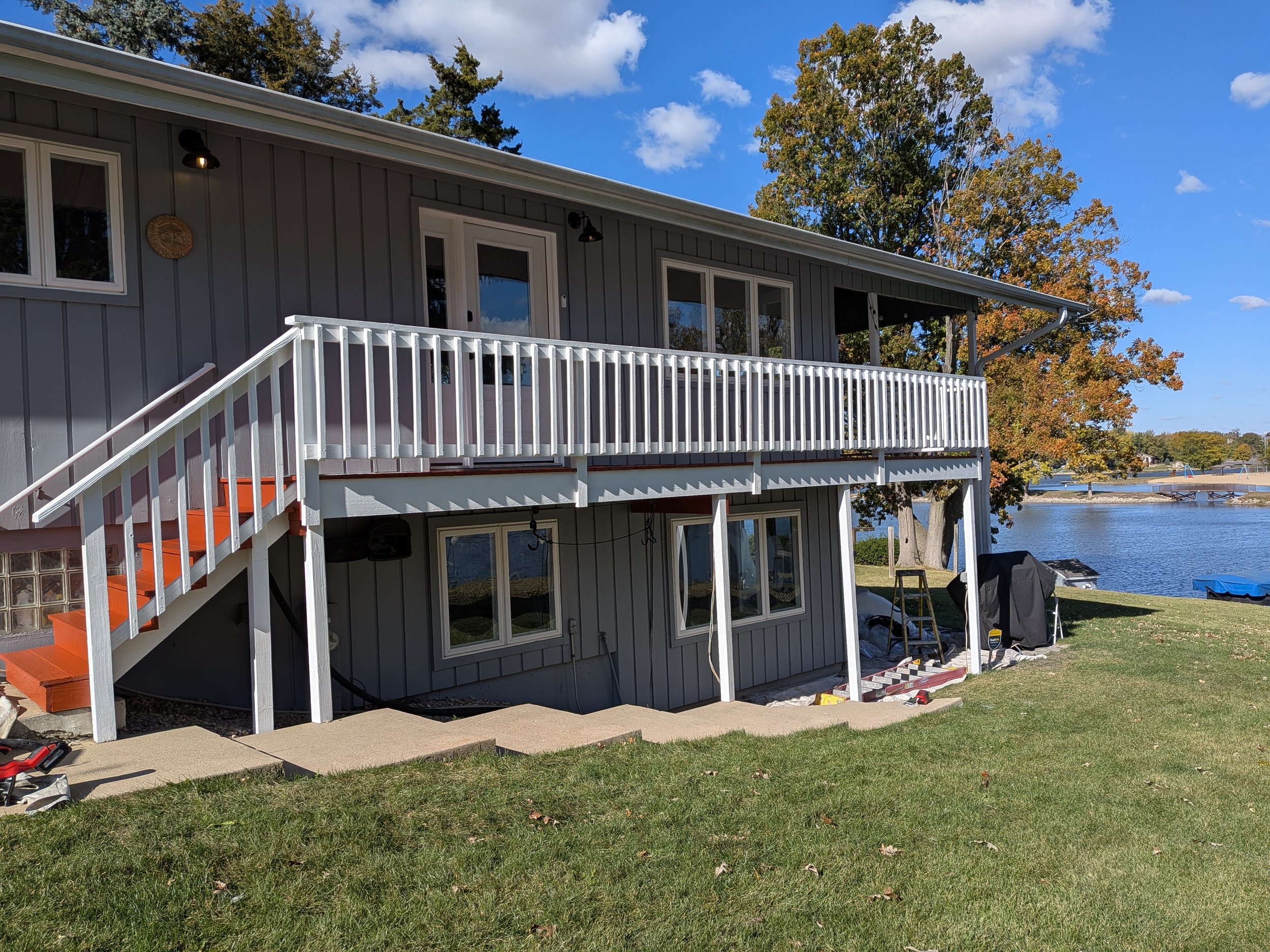 A two-story house with a gray exterior, white railing balcony, and a set of stairs with orange steps leading to the upper door. The house is near a lake with trees and a blue sky in the background.