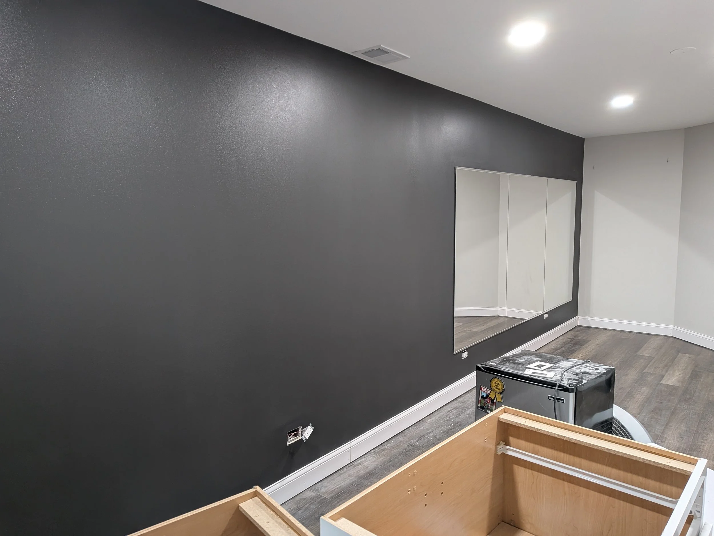 Empty room with a dark gray accent wall, a large mirror on the wall, hardwood floors, ceiling lights, a small cooling unit, and partially assembled furniture in the foreground.