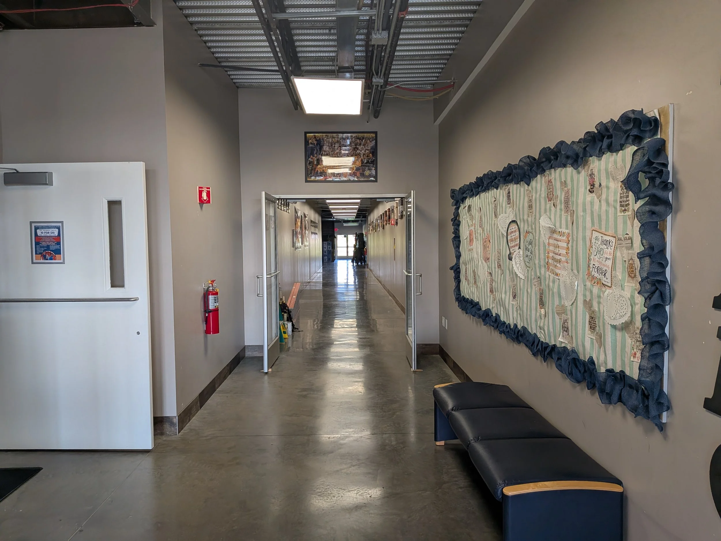 Indoor corridor with a bulletin board on the right wall, a bench beneath it, and a doorway ahead leading to a bright area. A fire extinguisher is mounted on the wall to the left.
