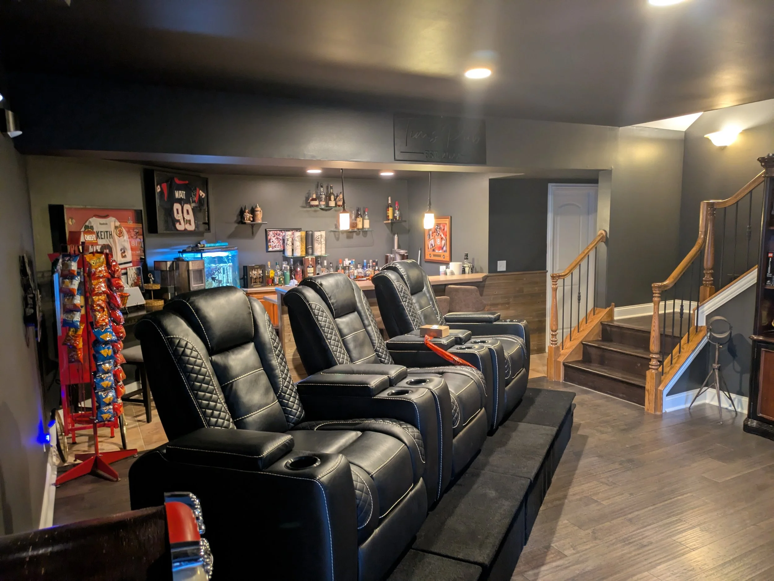 Home theater room with four black leather recliners, a bar area with liquor bottles, a fish tank, sports memorabilia, snack rack, and wooden stairs leading to another level.