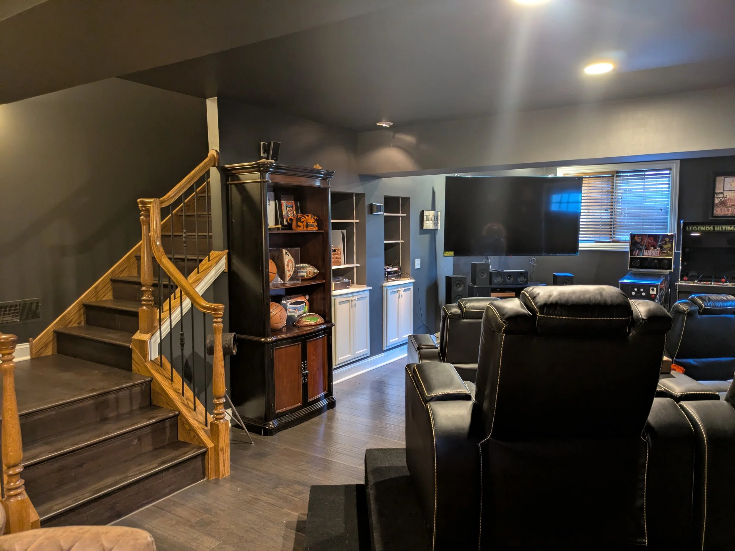 Living room with dark walls, leather recliners, large flat-screen TV, arcade game, and wooden stairs leading up. Shelves with sports memorabilia and a window with blinds are also visible.