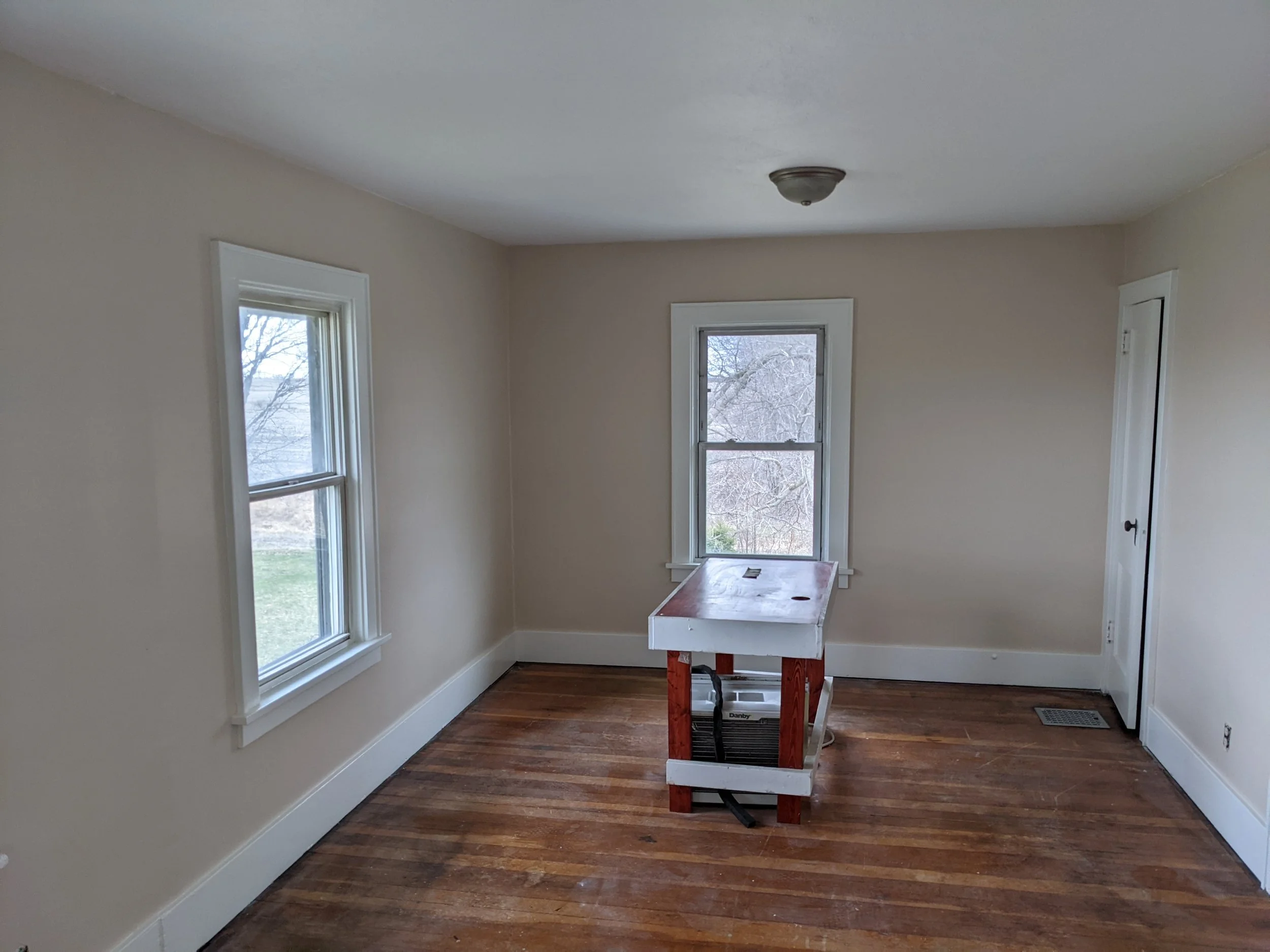 Empty room with hardwood floor, two windows, a simple ceiling light, and a small door. There is a white and red work table in the center.