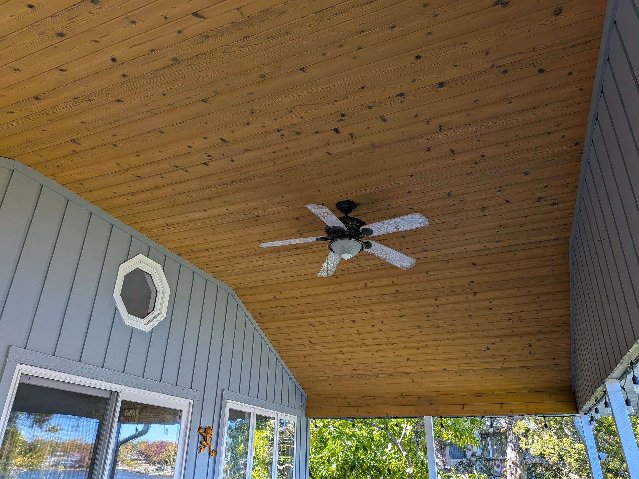 Covered porch area with a wooden ceiling, a white ceiling fan with four blades, a small octagonal window, windows with blinds, and trees outside.