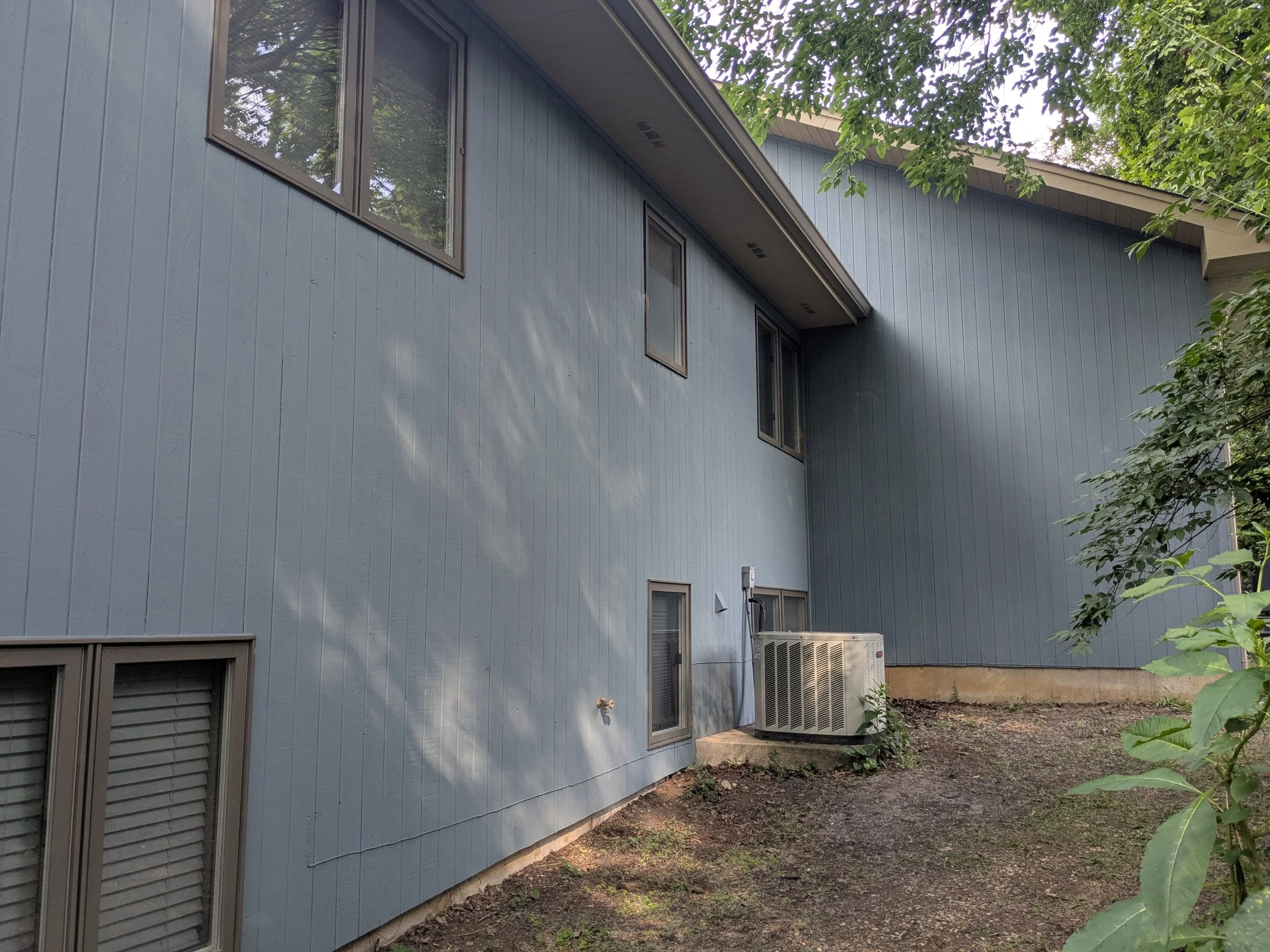 Rear exterior of a blue house with vertical siding, multiple windows, and an outdoor air conditioning unit on a concrete slab, surrounded by dirt and green foliage.