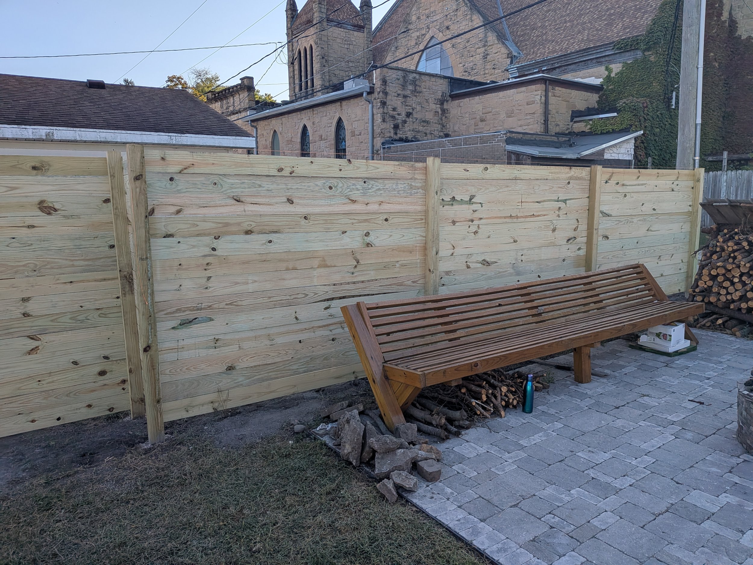 A wooden bench in front of a newly built wooden privacy fence on a paved outdoor patio area, with a church and trees in the background.
