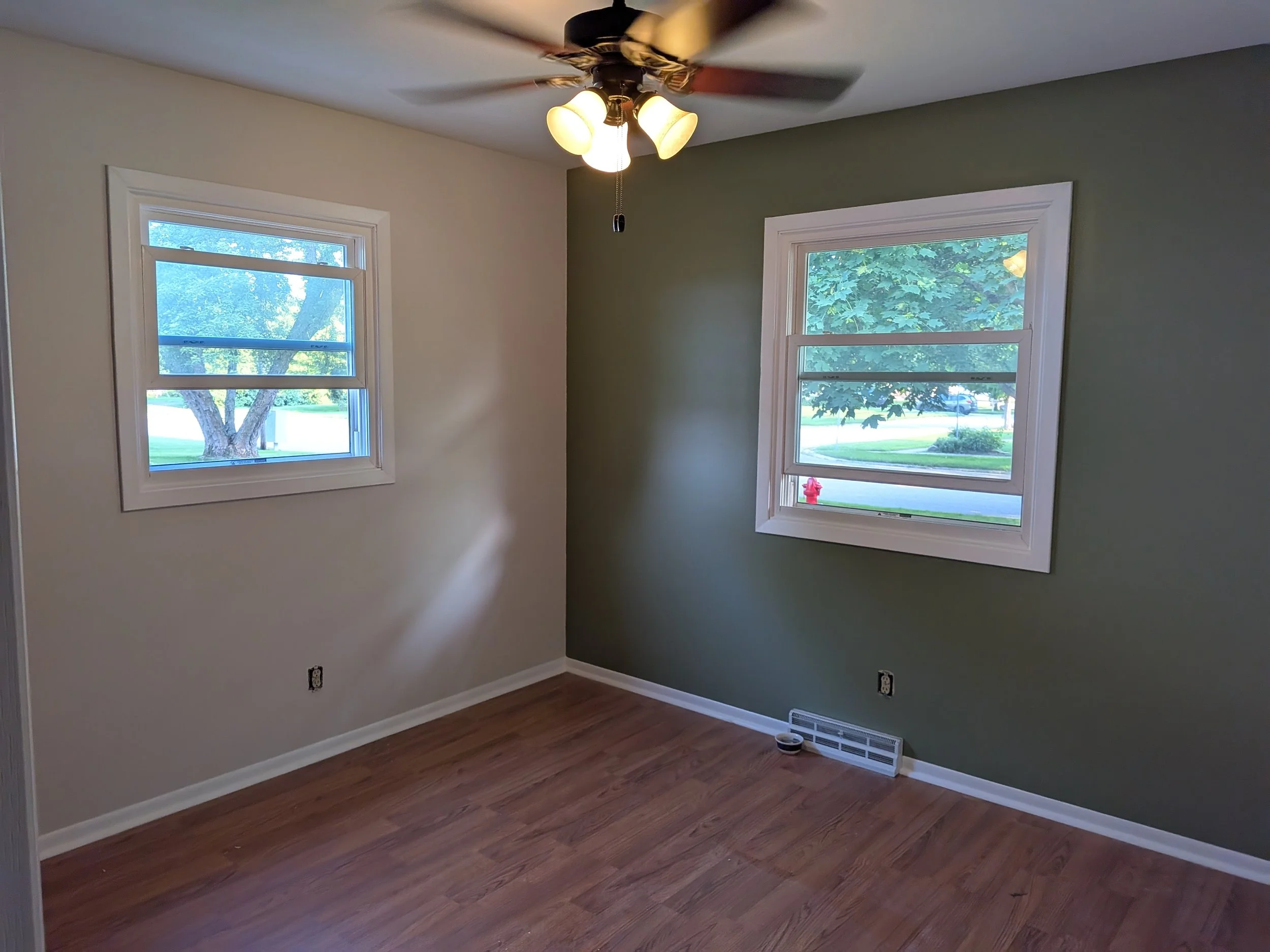 Empty room with two windows, ceiling fan with lights, hardwood floor, and a green accent wall.
