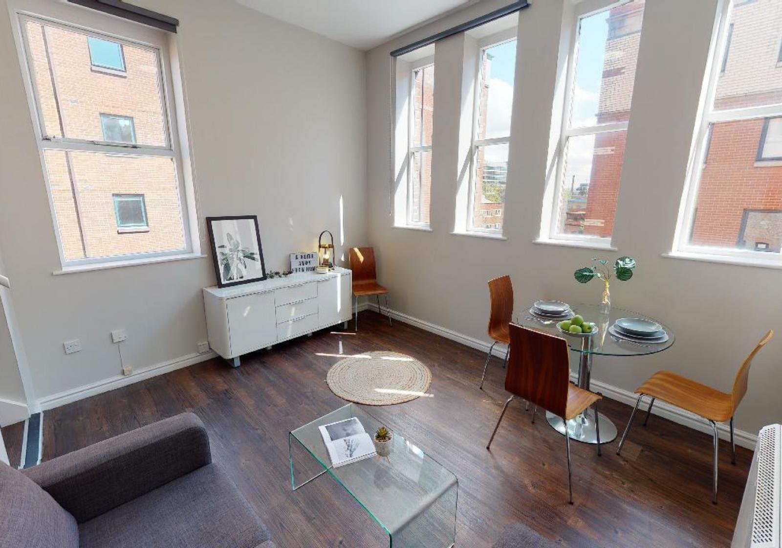 Modern living room with large windows, hardwood floors, glass coffee table, small round dining table with chairs, white sideboard, and neutral decor.