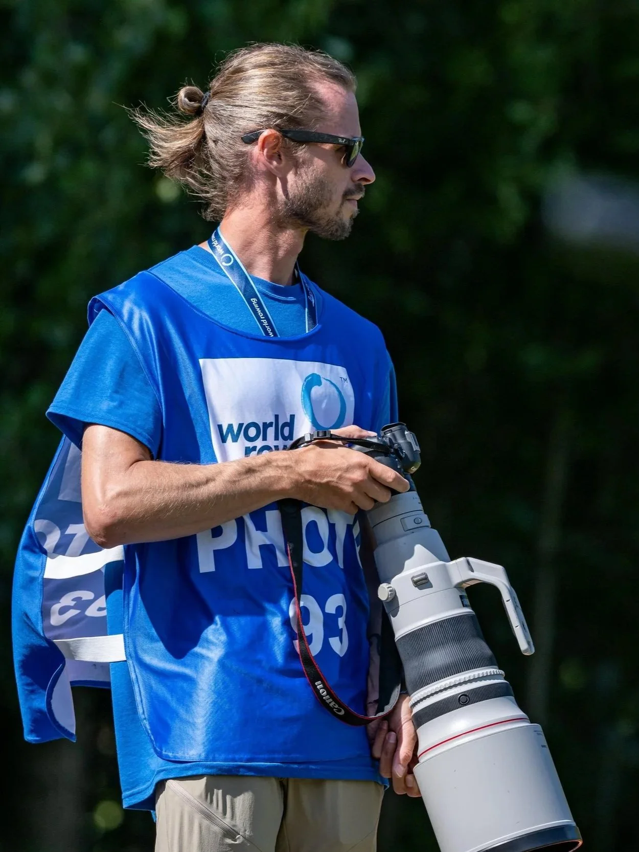 A man, Anthony Benoit, wearing a blue World Rowing vest holding a professional camera with a large telephoto lens, standing outdoors.