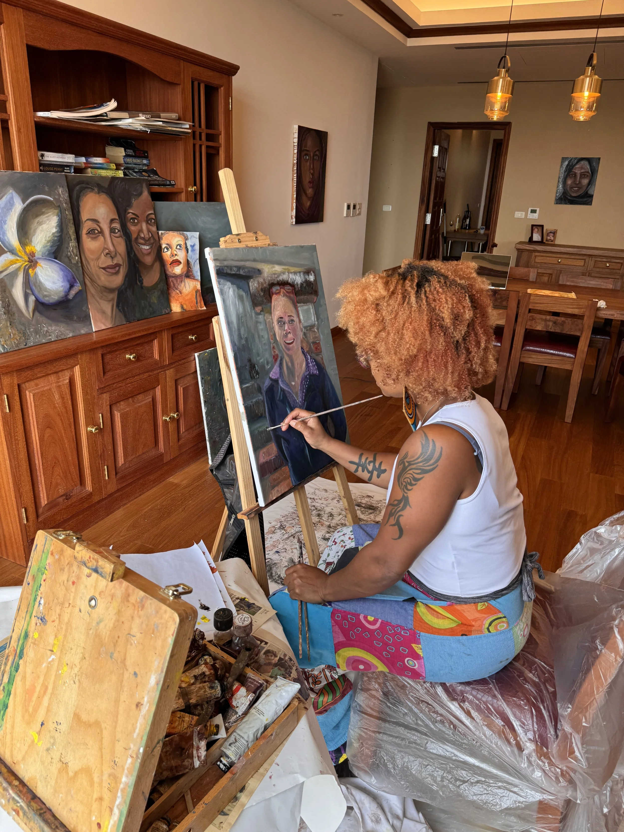A woman with curly hair and tattoos, sitting cross-legged on a plastic-covered chair, painting a portrait on a canvas in a home studio. Art supplies and other portraits on shelves and walls are visible in the background.