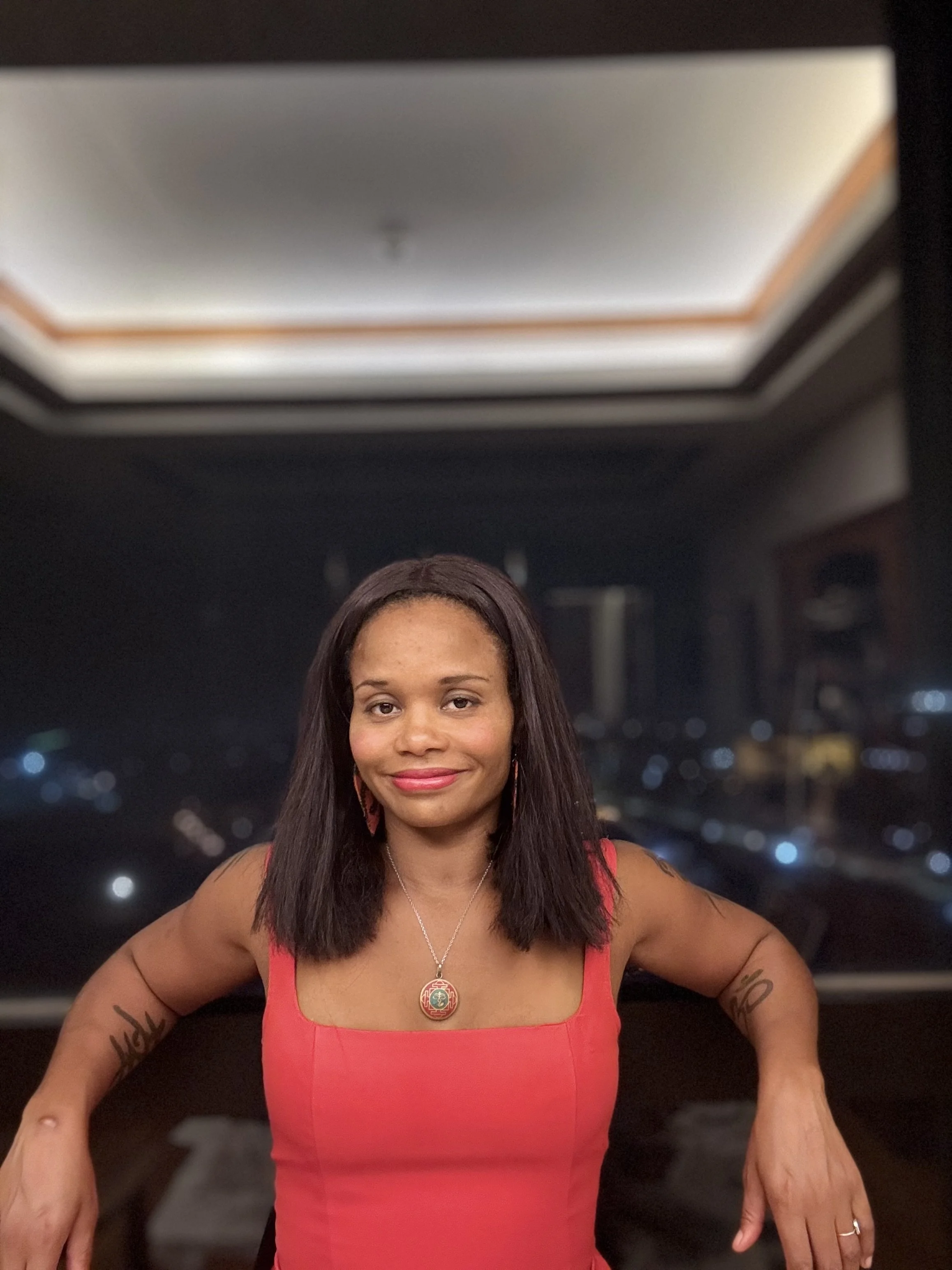 A woman with shoulder-length dark hair wearing a red sleeveless top and necklace, sitting in front of a large window at night with city lights in the background.