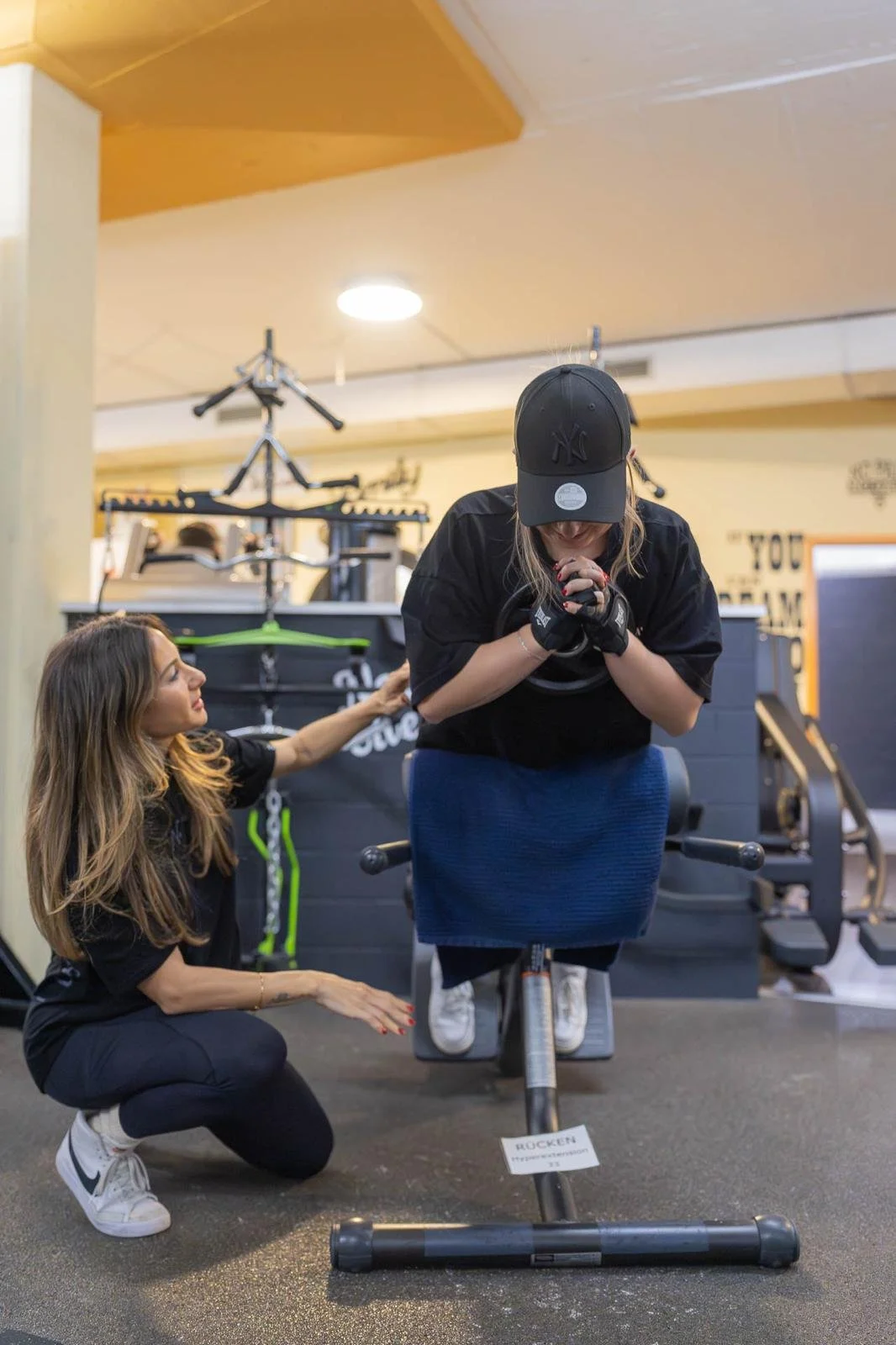 Eine Frau macht eine Übung auf einem Rückenstrecker im Fitnessstudio, eine andere Frau steht neben ihr und gibt Anweisungen.