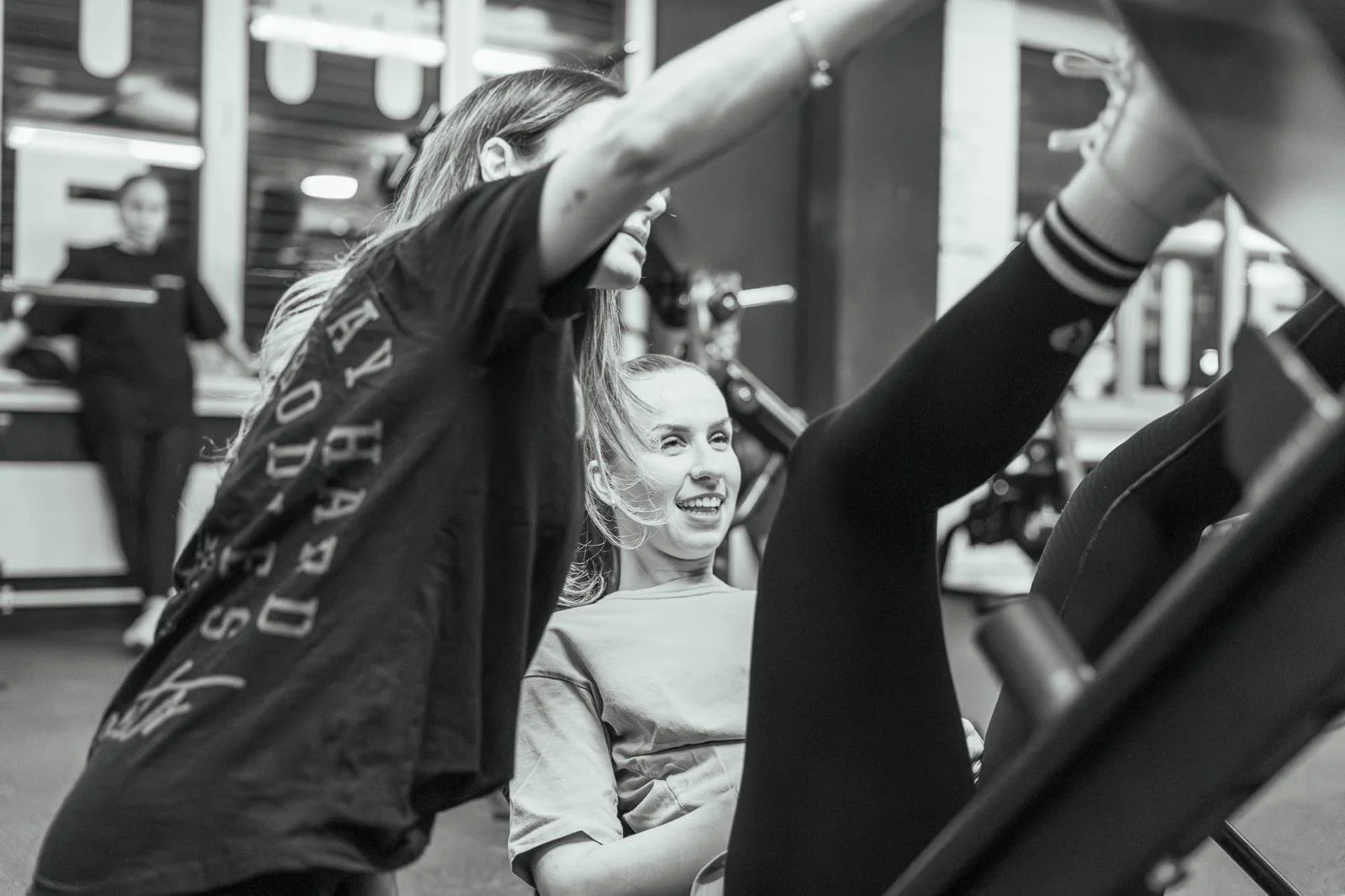 Zwei Frauen trainieren zusammen im Fitnessstudio, eine sitzt auf einem Gerät und lacht, die andere assistiert beim Training.