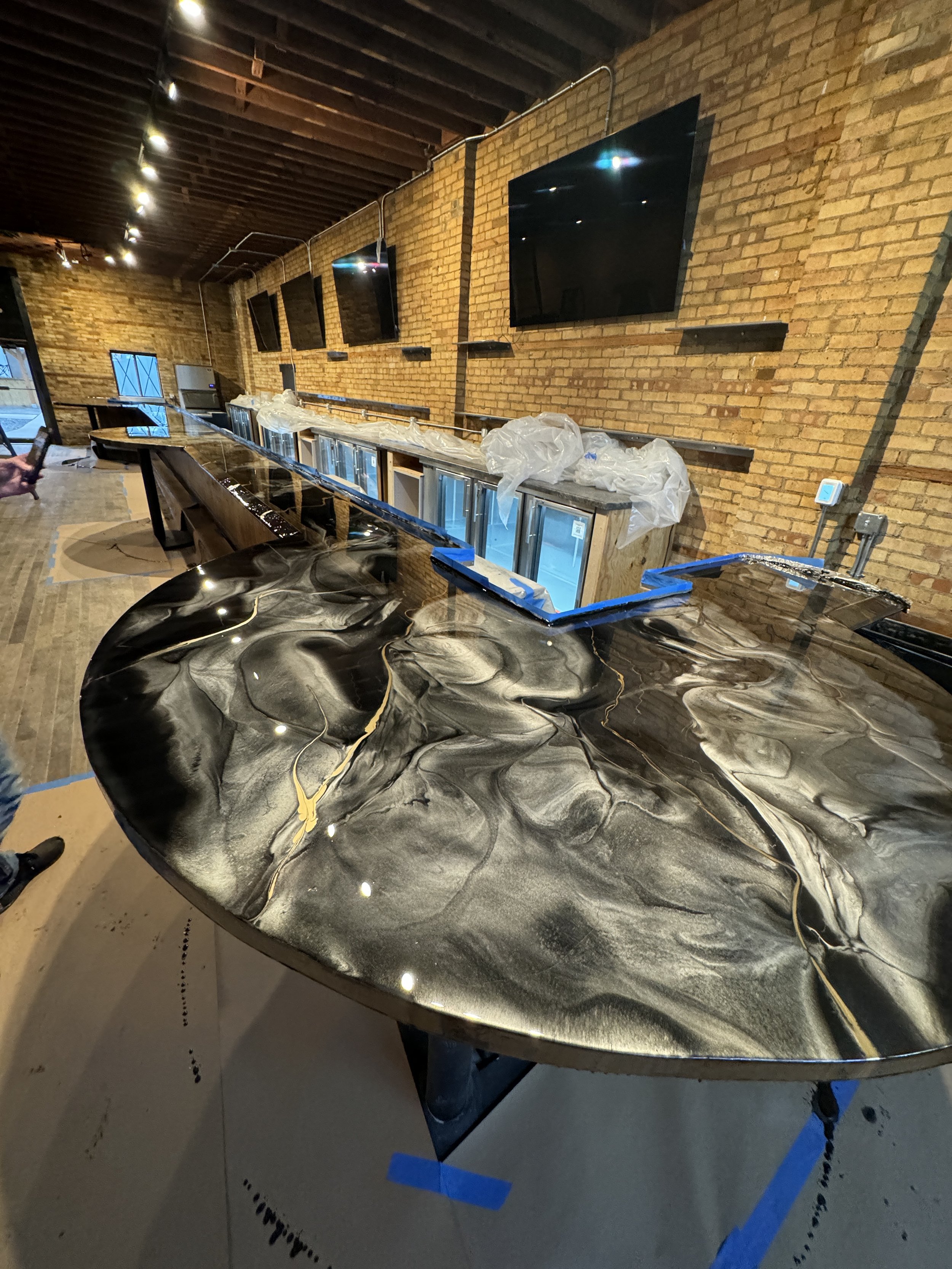 Interior of a modern bar with a marble countertop, exposed brick walls, multiple flat-screen TVs, and high ceilings with visible beams.