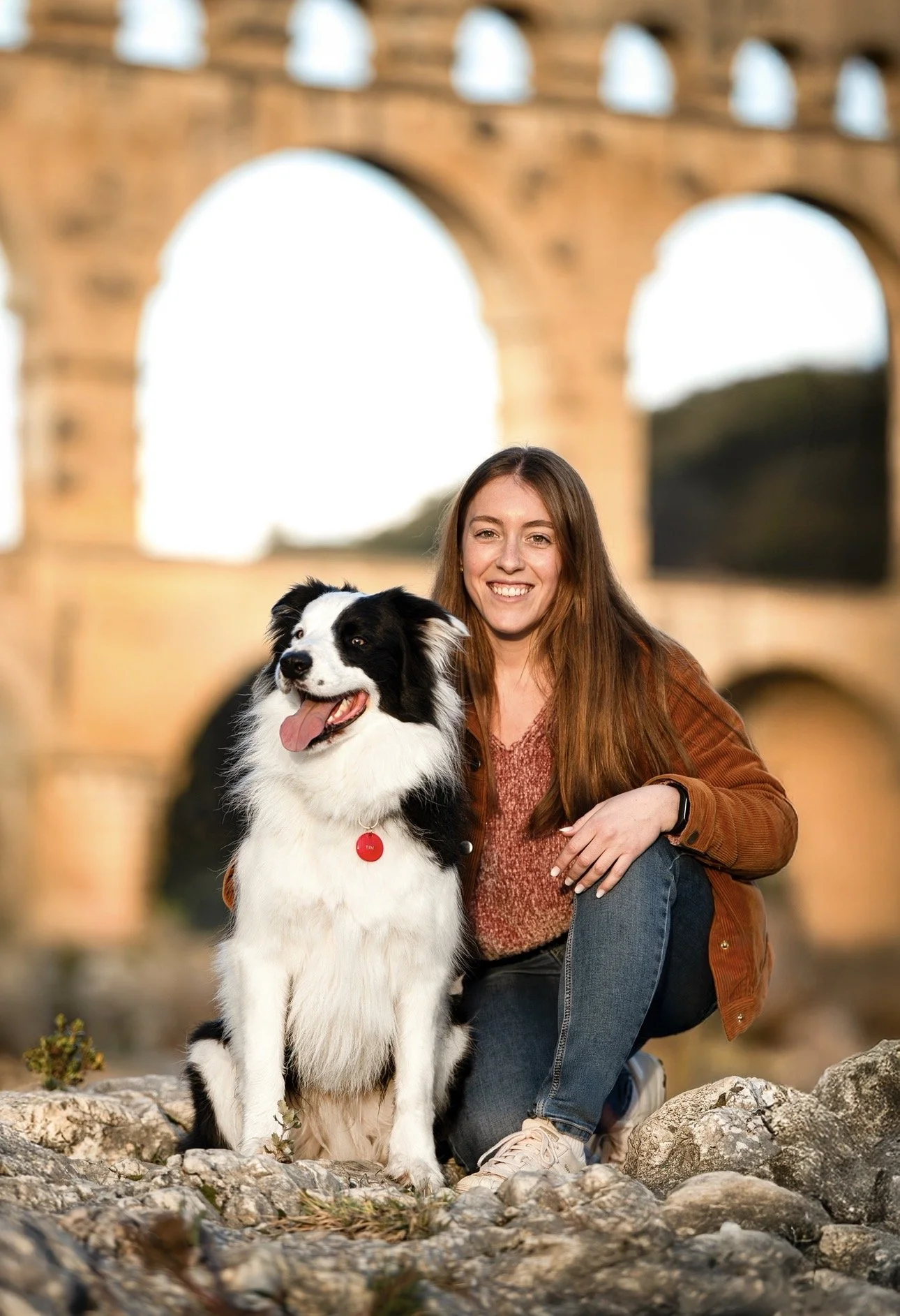 Une femme souriante avec un chien Border Collie noir et blanc devant l'arc de Triomphe de l'Aquila, le monument antique en pierre en arrière-plan.