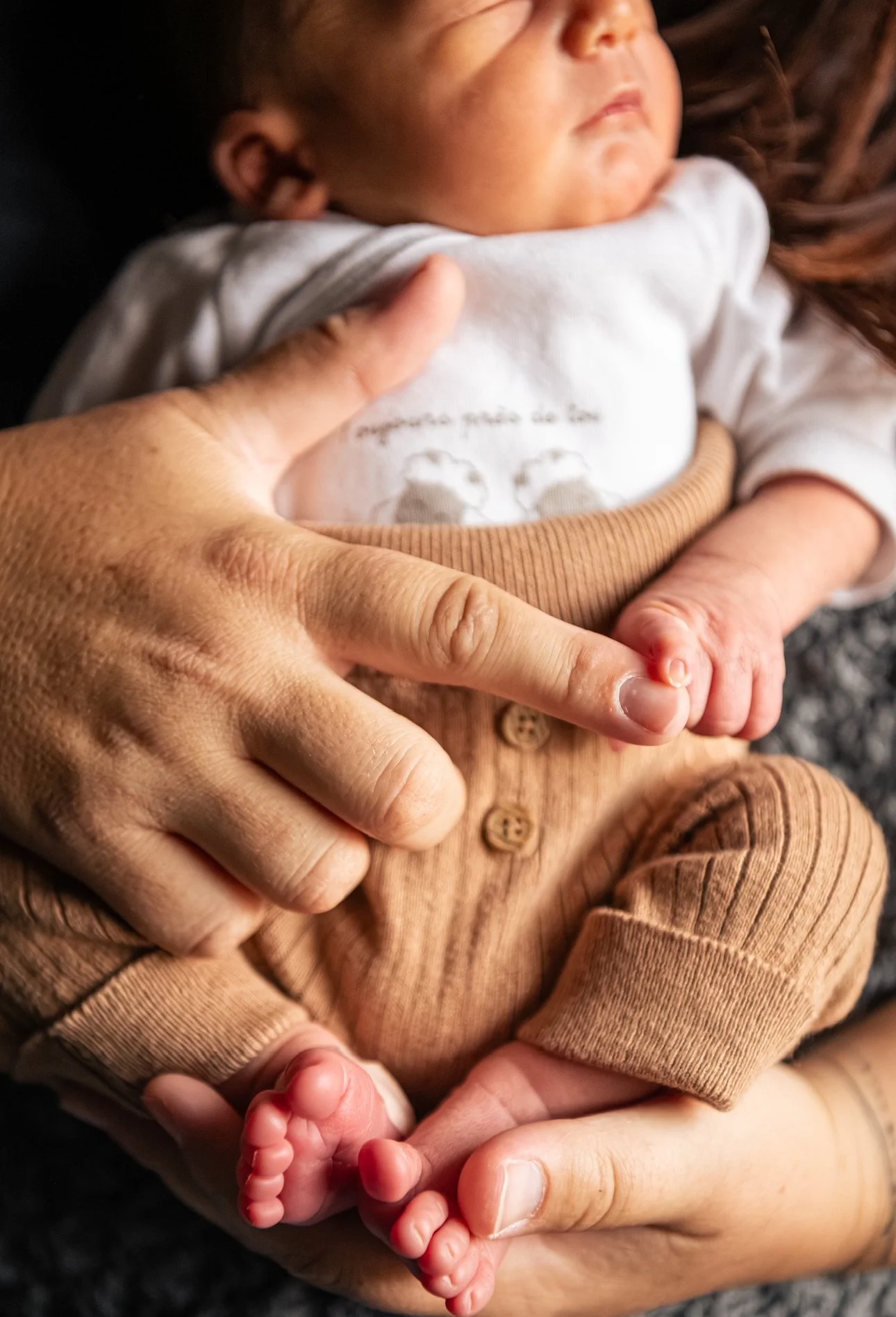 Un bébé allongé dans les bras d'une personne, vêtement beige, mains supportant délicatement le bébé, qui a de petits pieds nus.