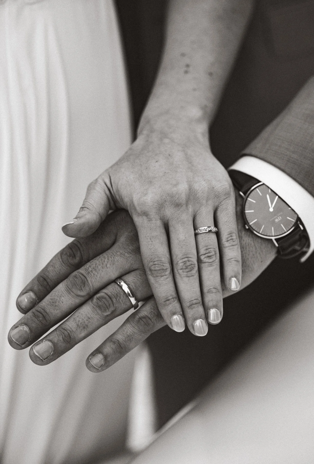 Une photo en noir et blanc montrant deux mains portant des alliances, une main humaine portant une montre élégante et un homme en costume visible partiellement avec une chemise blanche.