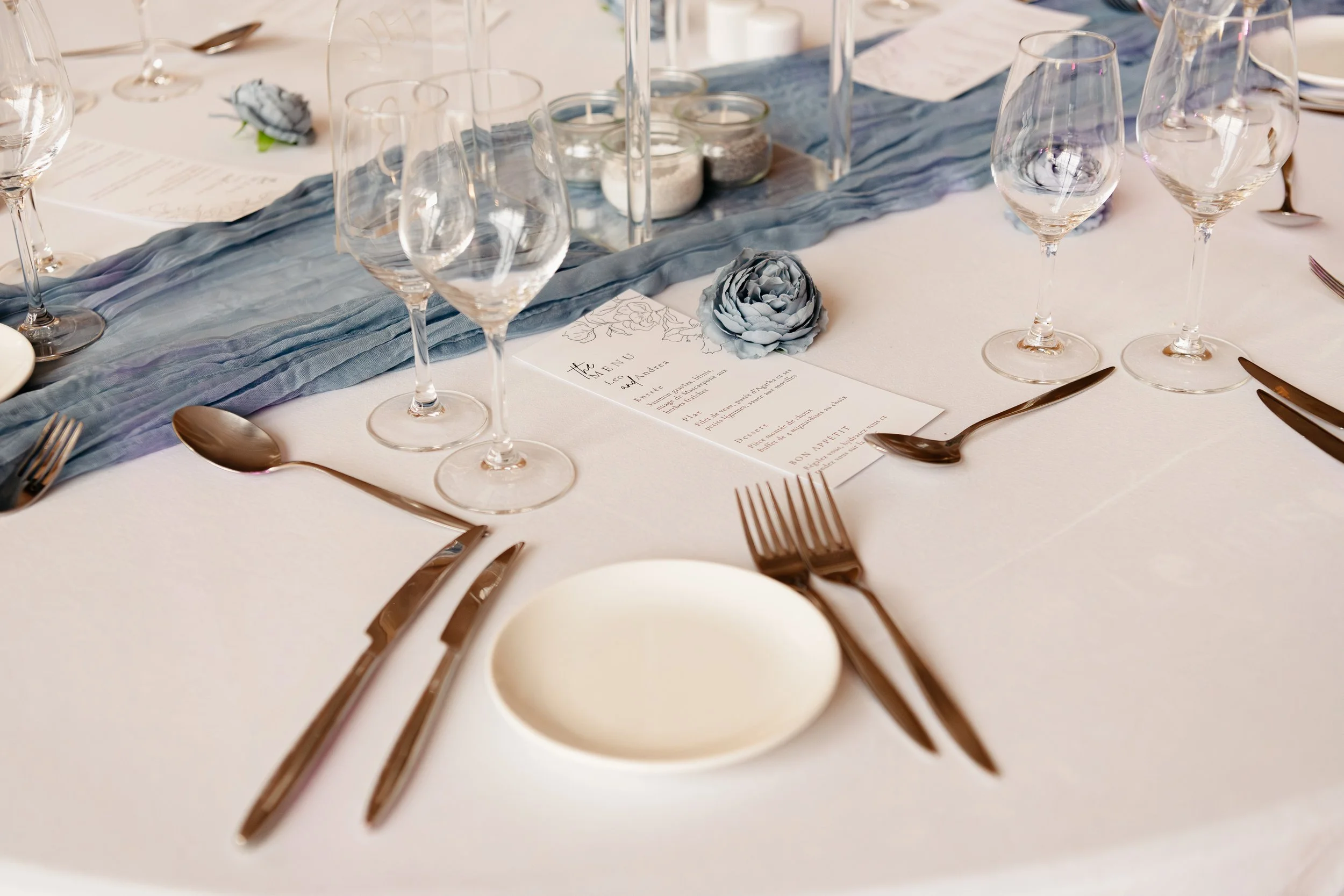 Table de mariage décorée avec des verres, des couverts en métal, une nappe blanche, et un centre de table avec un drapé en tissu bleu et des fleurs en papier bleu.