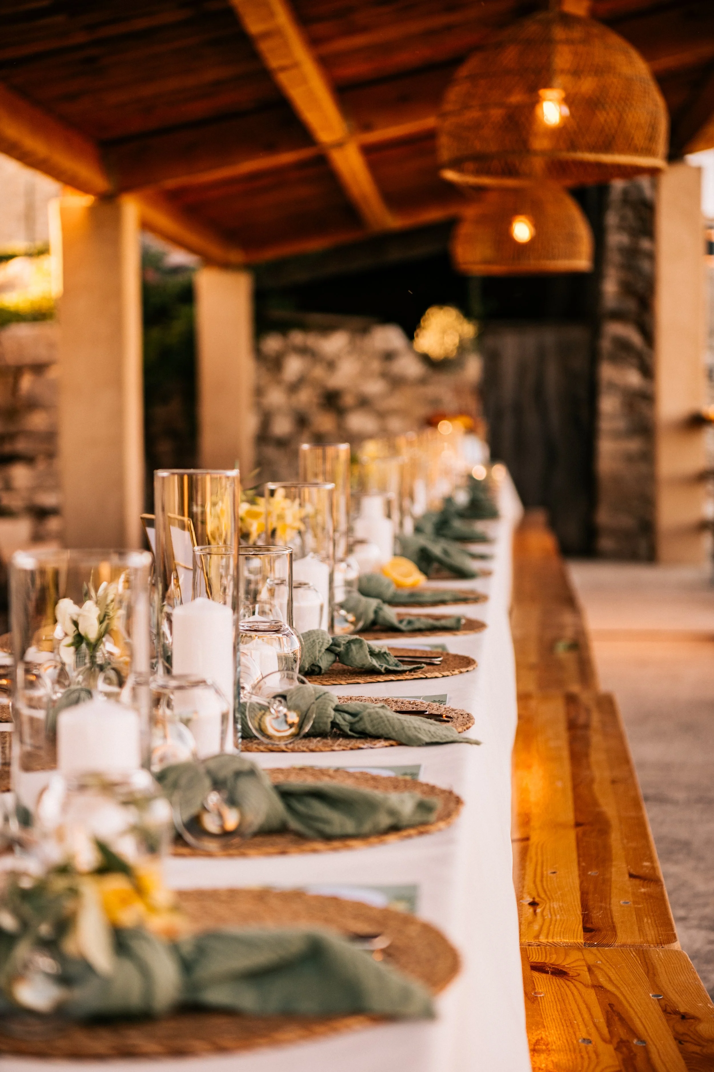 Table décorée pour un repas en plein air avec des bougies, des fleurs, et des place-settings, sous un toit en bois avec des lampes suspendues en osier.