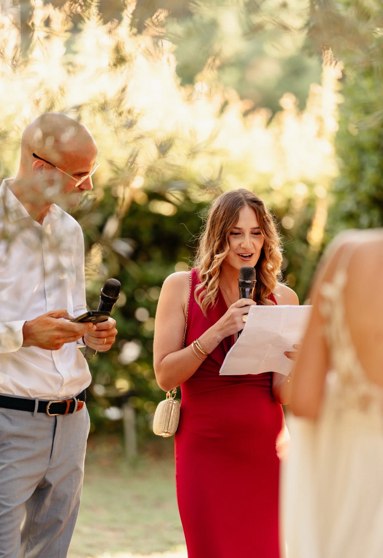 Une femme en robe rouge lit un discours lors d'un mariage en plein air, un homme à côté d'elle consulte un téléphone. Autour, la nature et la lumière du soleil créent une ambiance chaleureuse.