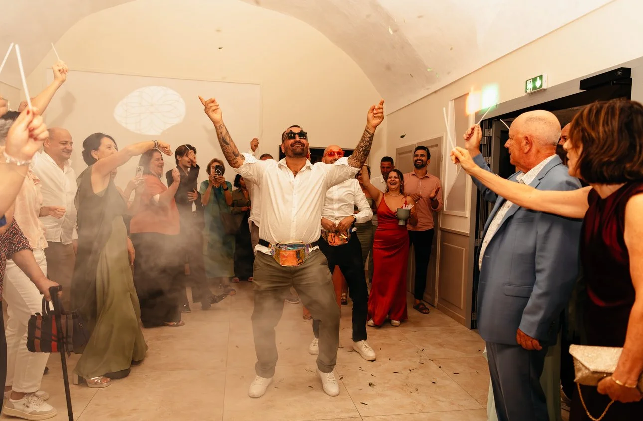 Un groupe de personnes célébrant lors d'une fête, avec un homme au centre dansant avec des bras levés, entouré par d'autres qui lèvent des baguettes brillantes ou fidèle dans une ambiance joyeuse.