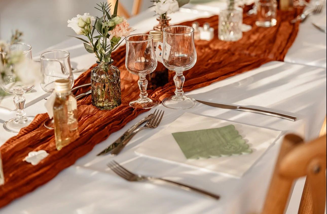 Table dressée pour un dîner avec une nappe blanche, une chemin de table en tissu marron, assiettes, verres à vin, bouquets de fleurs et serviettes en tissu vert.