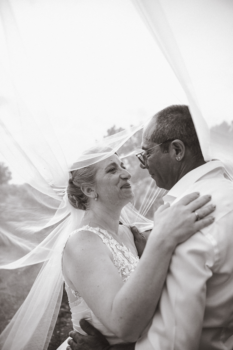 Un couple de mariés danse sous un voile de mariée lors de leur mariage en plein air, en noir et blanc.