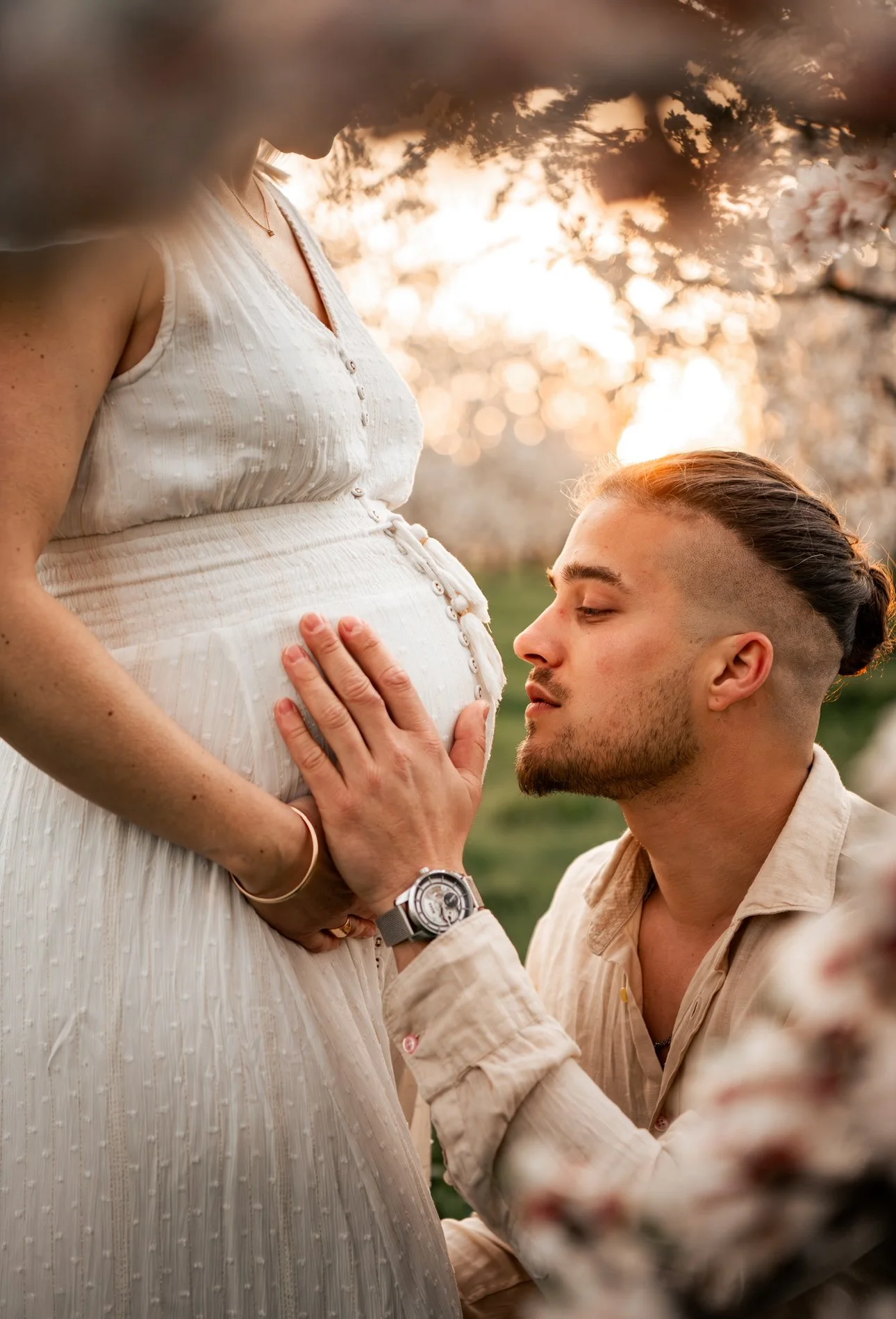 Un couple lors d'une séance photo avec une femme enceinte et un homme, dans un cadre extérieur avec des fleurs et un coucher de soleil.
