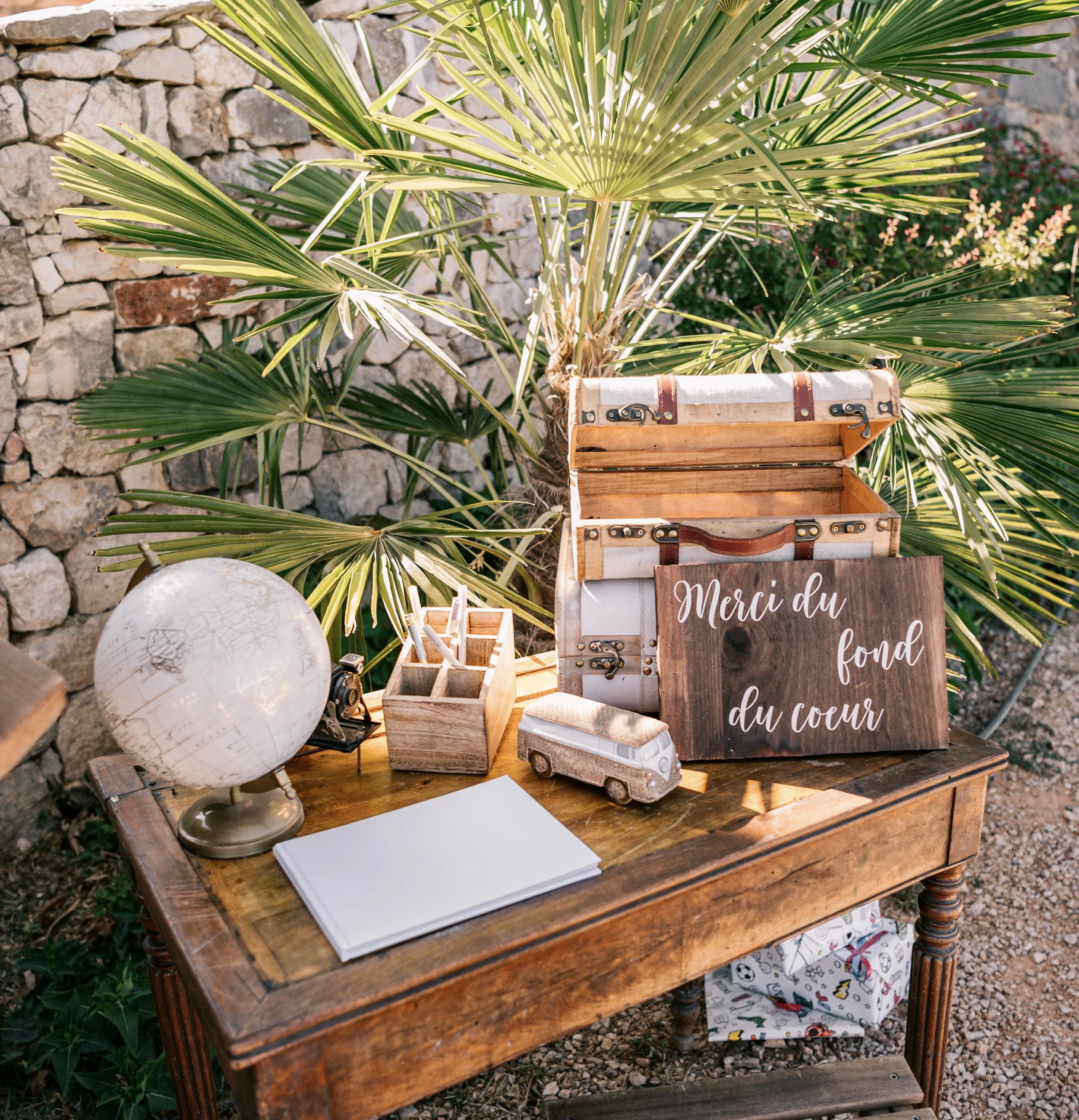 Un bureau en bois avec une globe, un petit camion en bois, un bloc-notes blanc et un contenant à crayons, devant une plante verte luxuriante et une pierre ancienne. Une boîte en bois ouverte et une affiche en bois avec le message 'Merci du fond du cœ