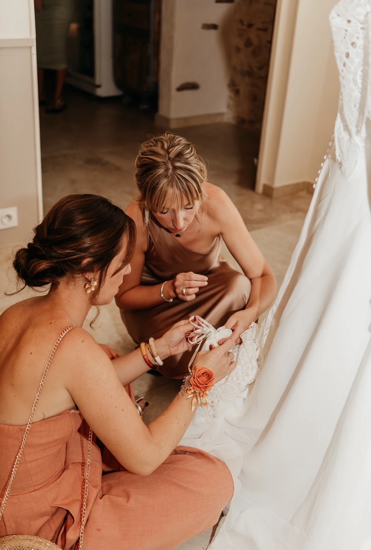 Deux femmes examinent une robe de mariage blanche, la femme à gauche porte une robe orange et un bracelet avec une fleur orange, la femme à droite porte une robe bronze et un bracelet. La scène se déroule dans une pièce intérieure.