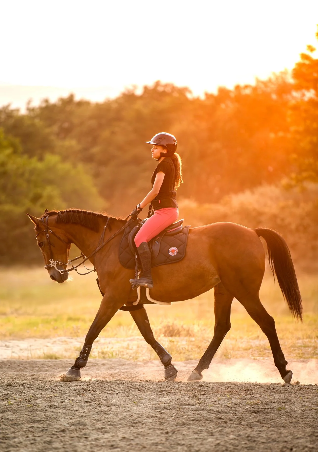 Une fille à cheval lors d'une promenade au coucher du soleil, portant un casque, un t-shirt noir, des pantalons roses et des bottes, en pleine nature.