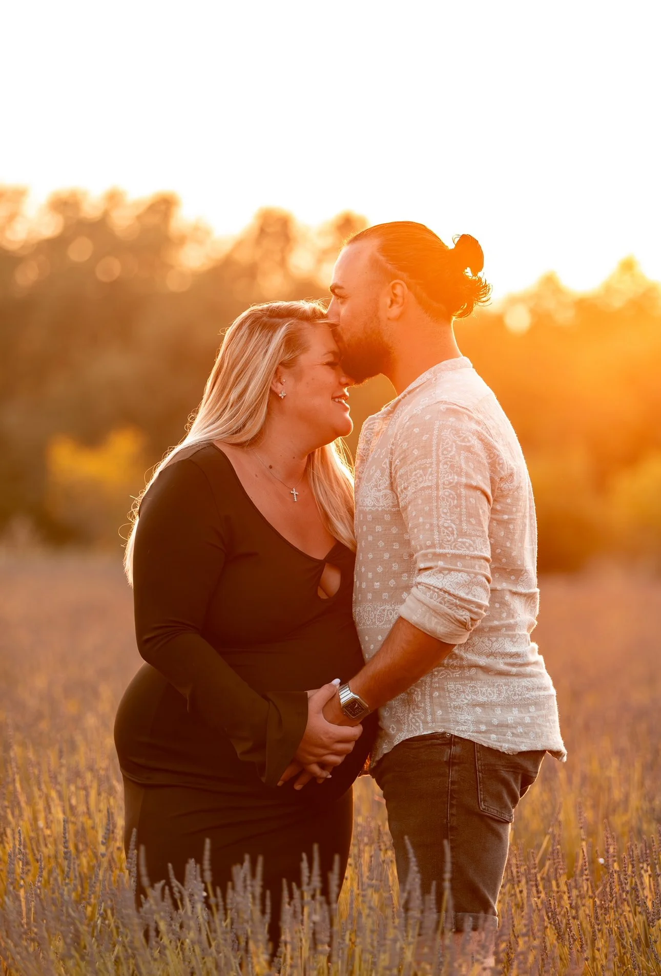 Un couple se tient dans un champ lors d'un coucher de soleil, partageant un moment tendre, l'homme embrassant la femme sur le front.