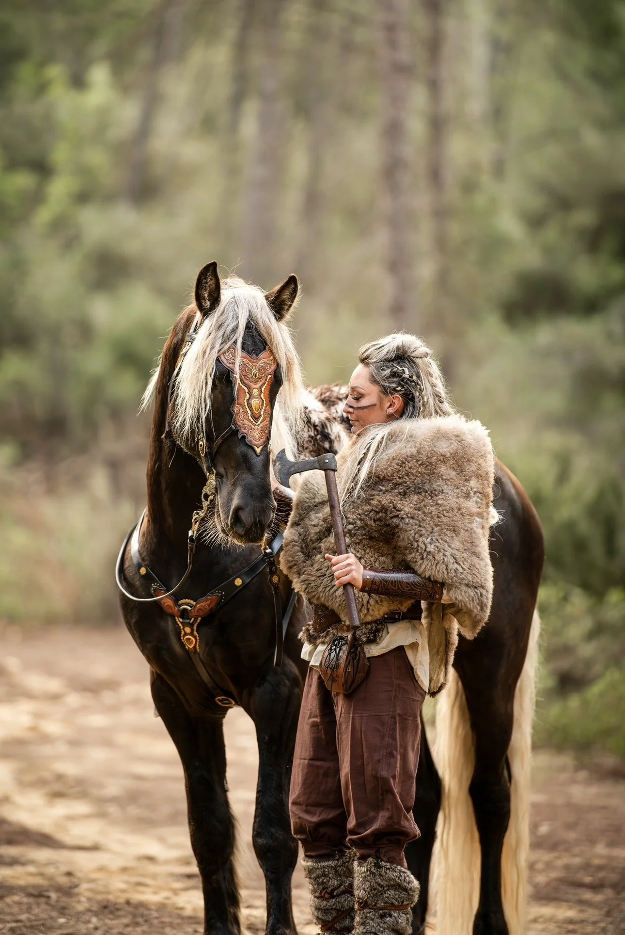 Une femme avec des cheveux gris épaisses en habits médiévaux en pelage, tenant une hache, face à un cheval noir et blanc dans une forêt.
