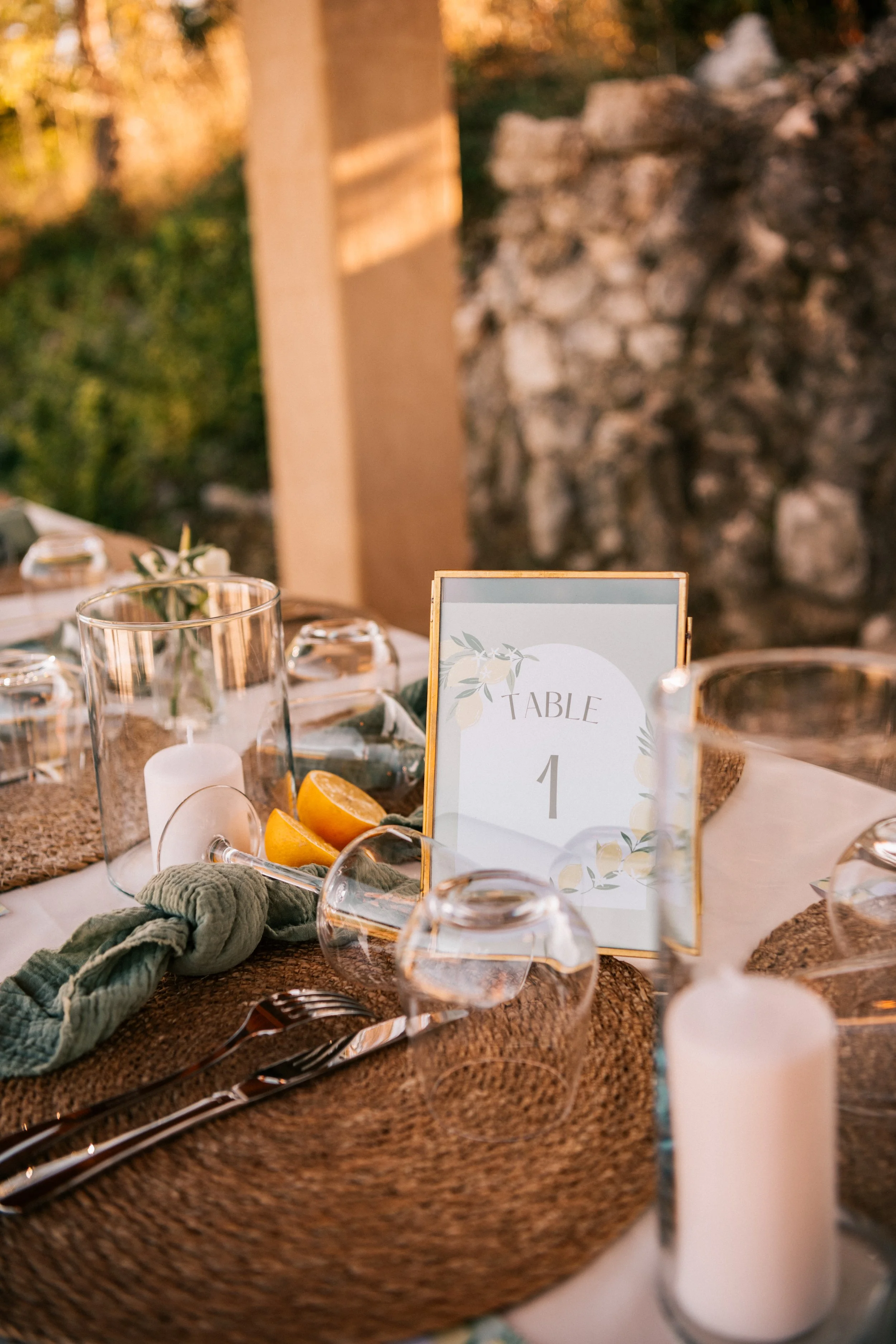 Table de dîner élégante avec des verres à vin, bougies, quartiers de citron, et un signet 'Table 1' dans un cadre doré, décorée avec un style champêtre et une ambiance chaleureuse en extérieur.