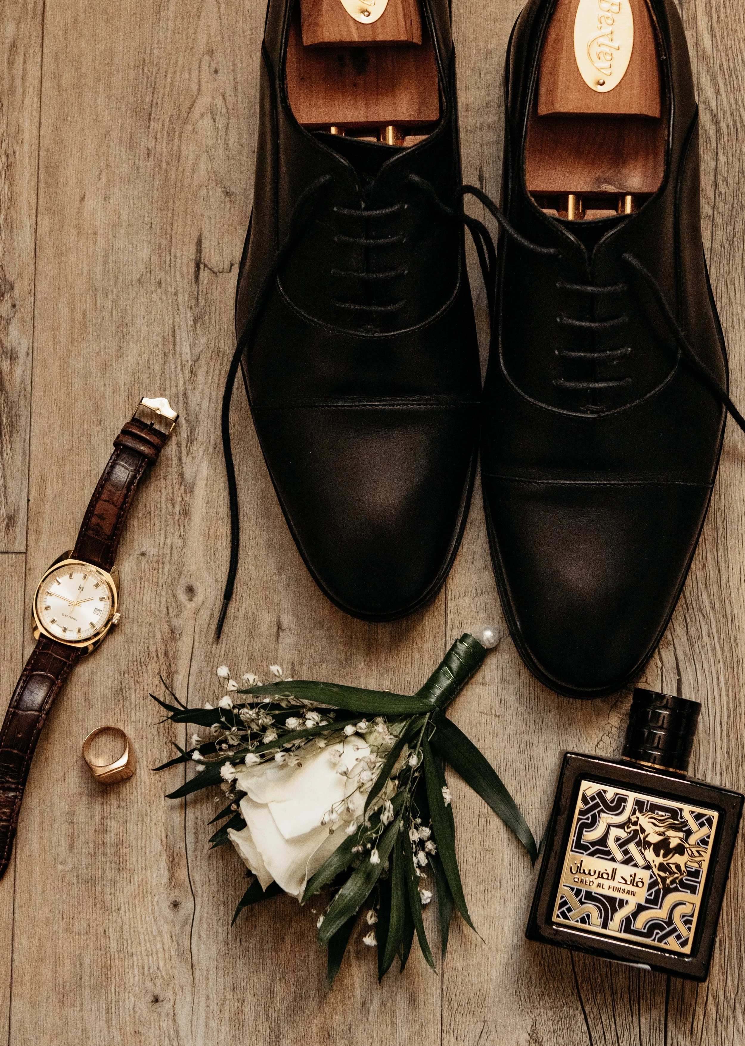 Chaussures en cuir noir avec semelles en bois, montre à bracelet en cuir marron, bague en or, bouquet de fleurs blanches, parfum en bouteille noire avec dessin de lion, et un gros anneau en or.