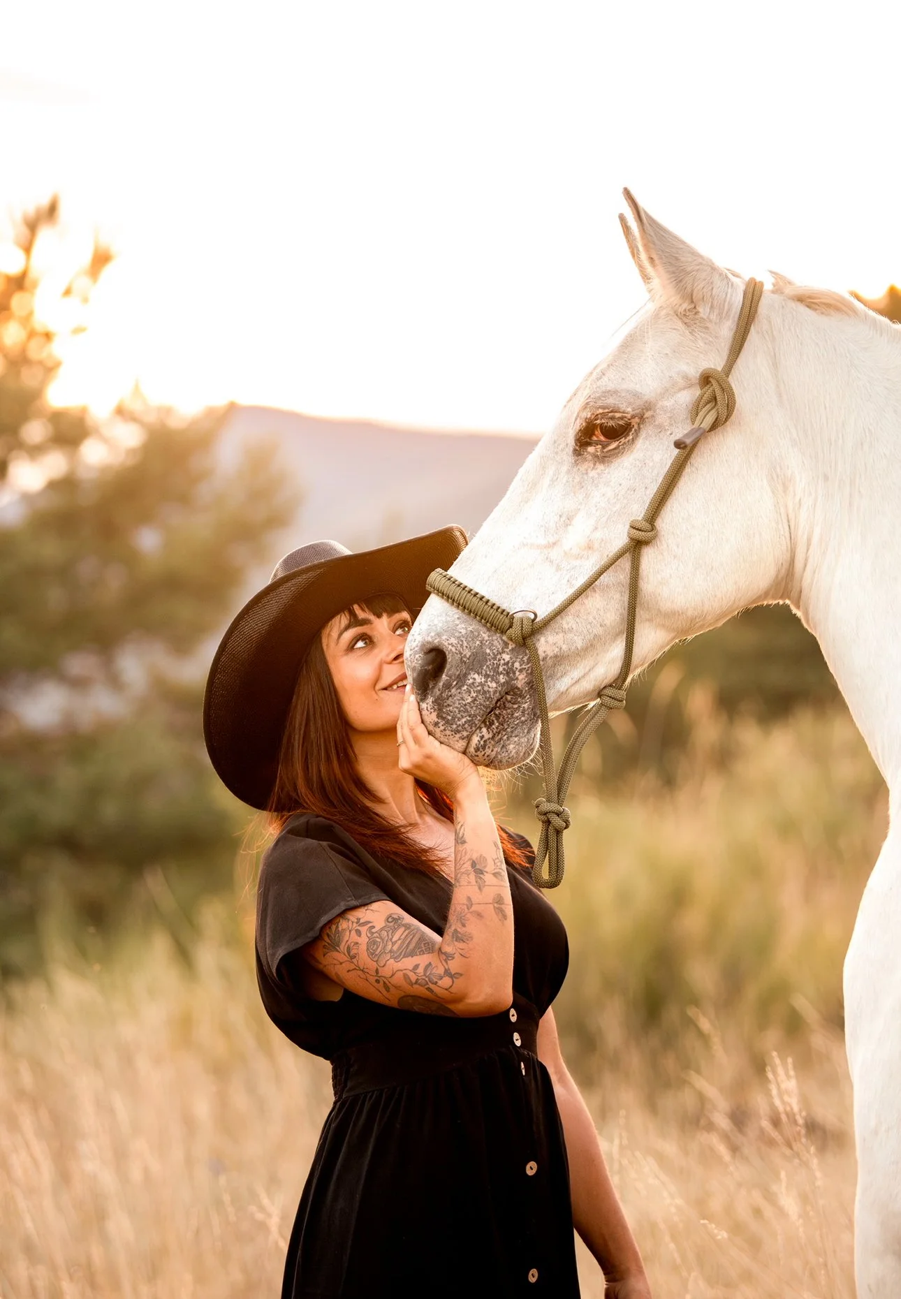 Une femme avec des tatouages portant un chapeau noir et une robe noire, caresse un cheval blanc dans un champ au coucher du soleil.