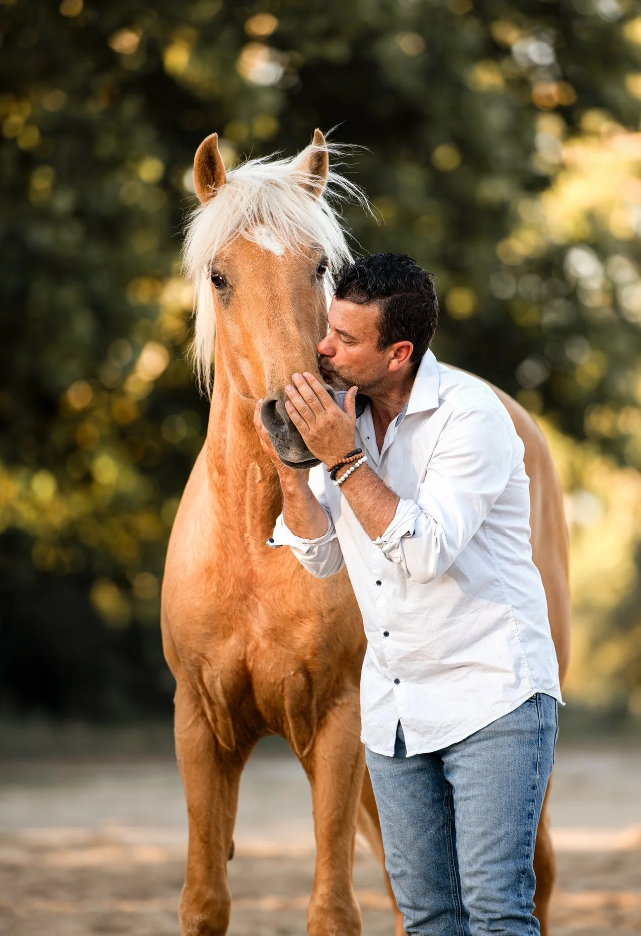 Un homme embrasse tendrement un cheval beige dans un environnement naturel flou avec des arbres en arrière-plan.