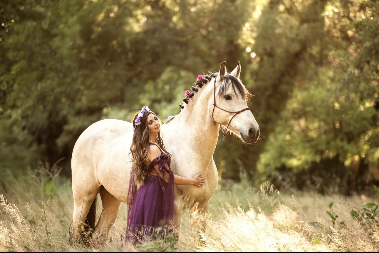 Une femme en robe violette avec une couronne de fleurs se tient près d'un cheval beige dans une forêt ensoleillée.