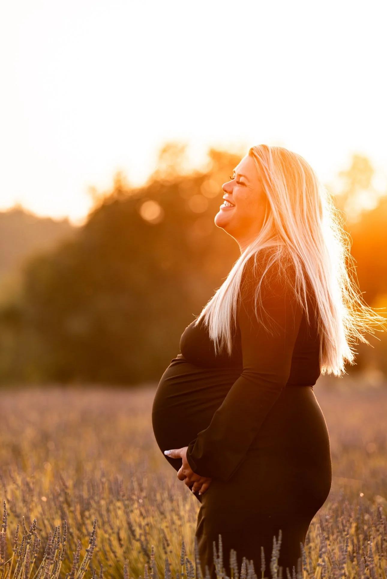 Une femme enceinte souriante avec des cheveux blonds longs, portant une robe noire, se tenant dans un champ de lavande lors du coucher du soleil.