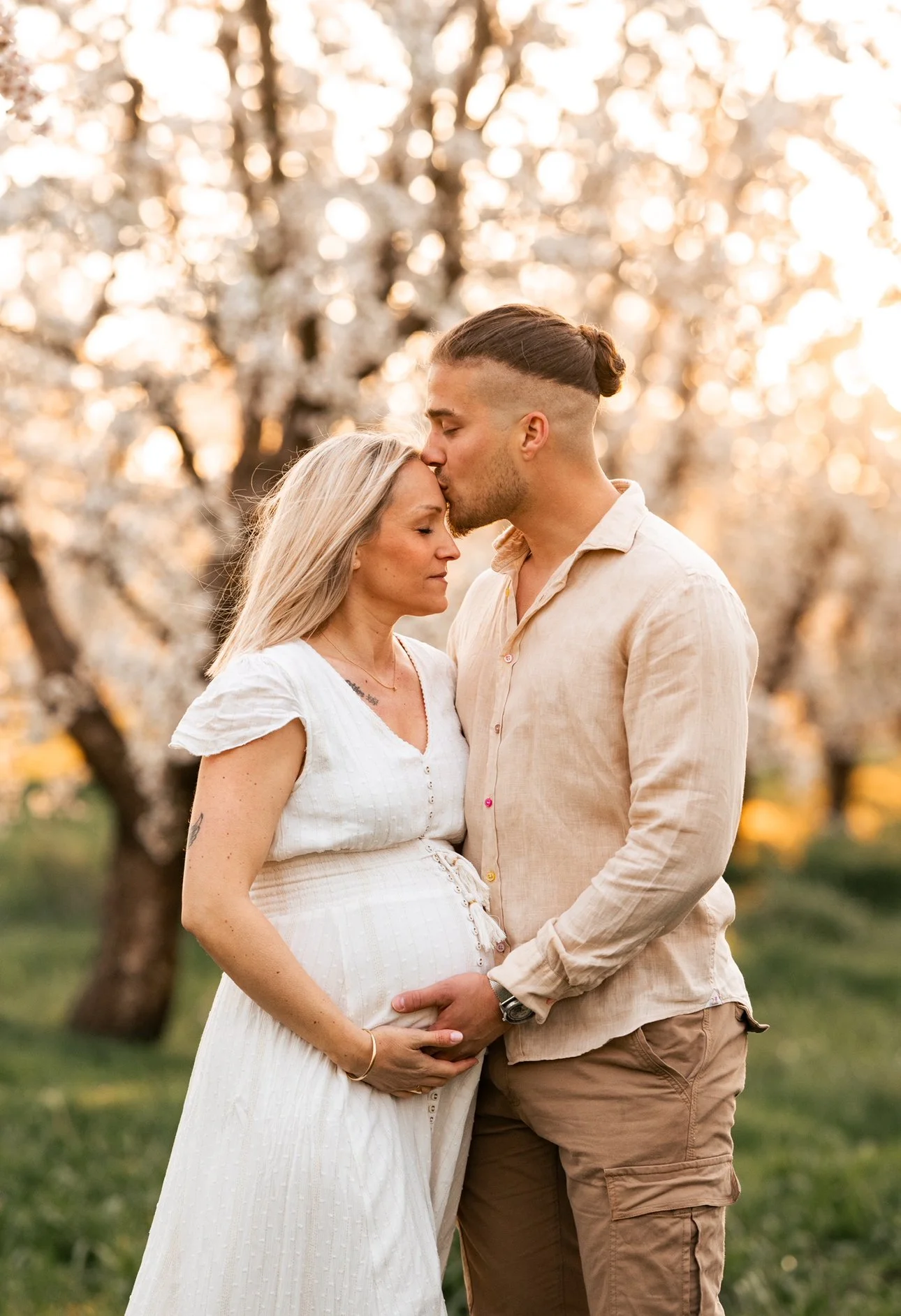 Un couple en grossesse, la femme enceinte porte une robe blanche et le couple se tient dans un parc avec des arbres en fleurs, durant un coucher de soleil.