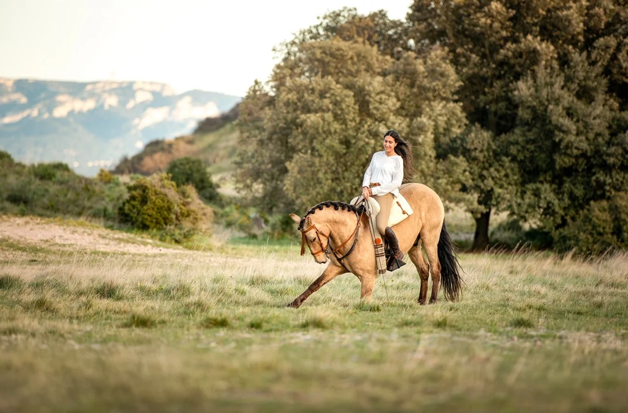 Femme à cheval dans un champ herbeux avec arbres et montagnes en arrière-plan.